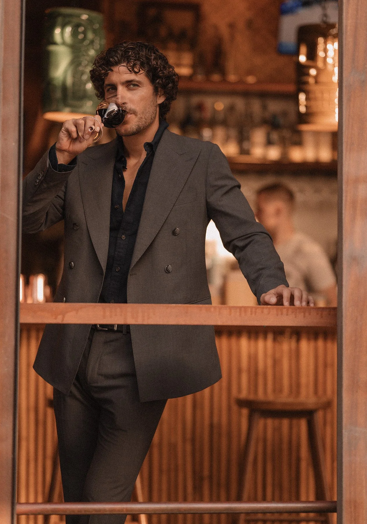 A man with curly hair, dressed in a dark suit jacket over a black shirt, is sipping red wine in a cozy, warmly lit bar or restaurant with wooden decor and a bartender or staff member in the background.