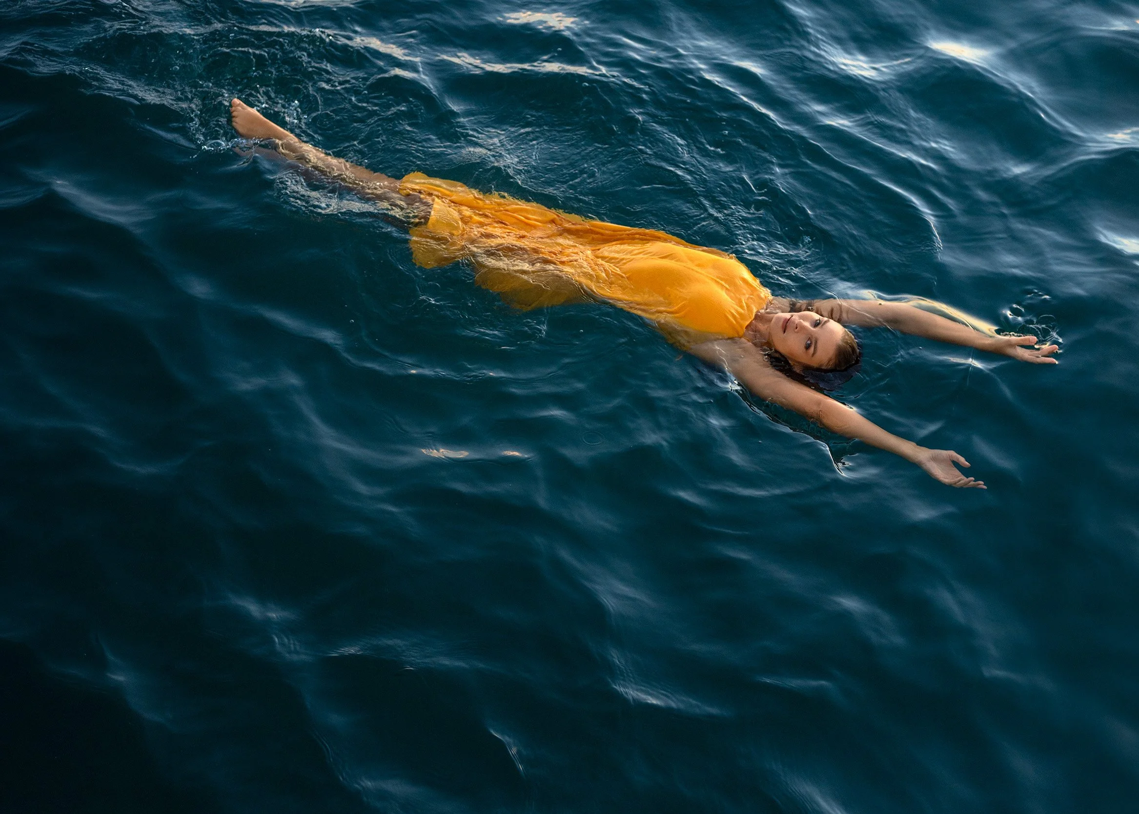 A woman in a yellow dress floating on her back in the ocean with arms outstretched and head tilted to the side.