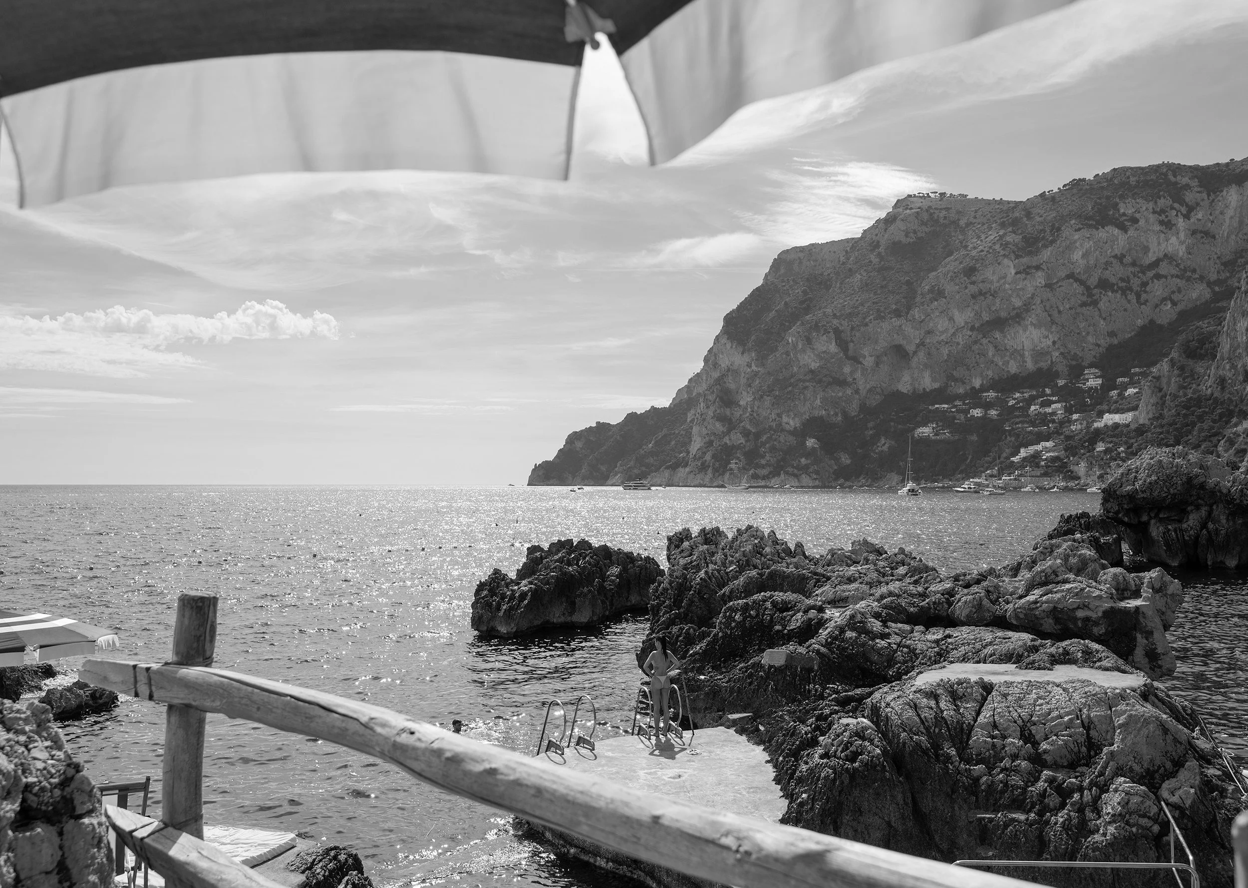 A black and white photo of a coastal scene with rocky shoreline, boats in the water, and cliffs with houses on them in the distance.