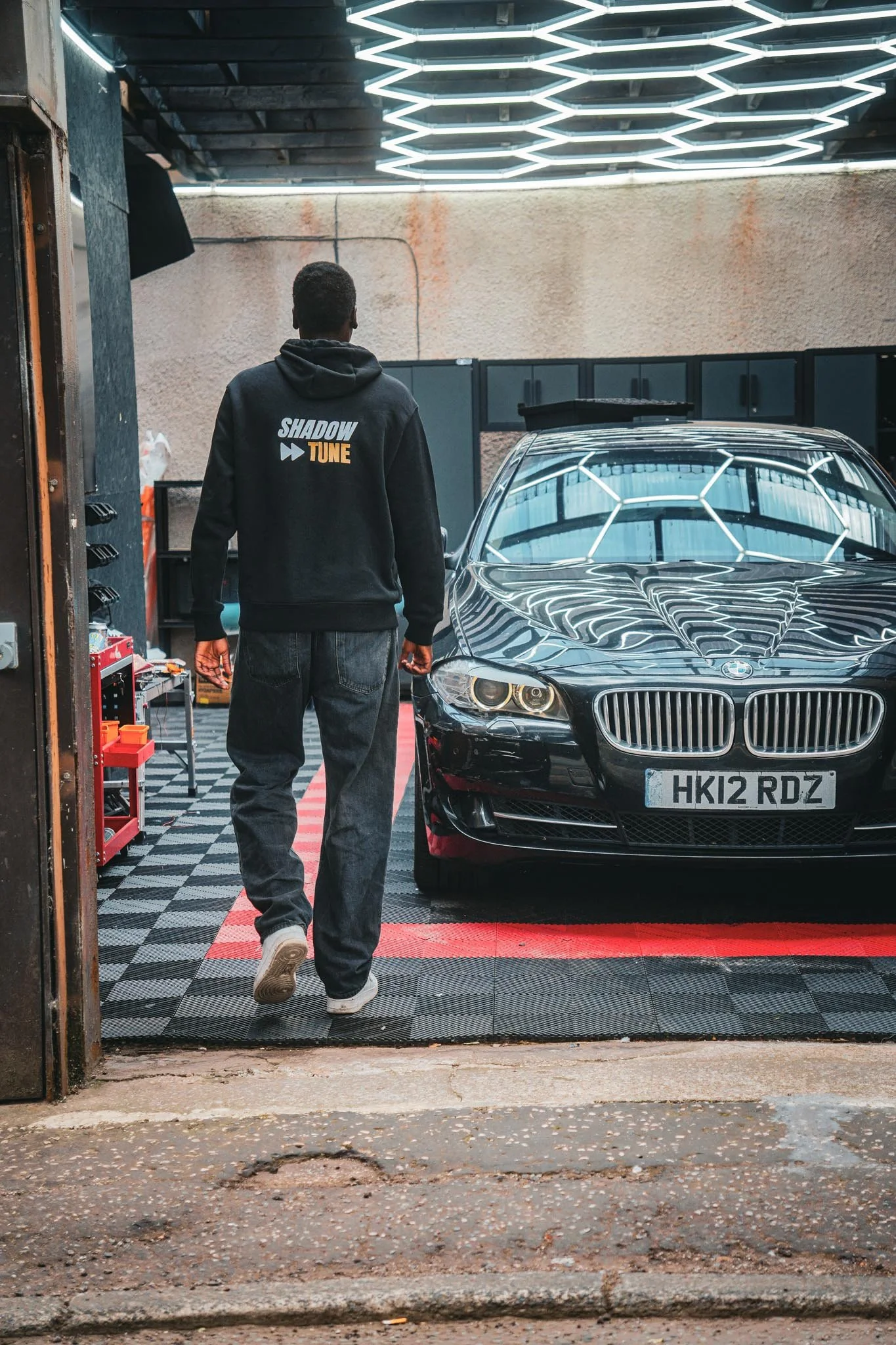 A person walking toward a black BMW car inside a garage with bright hexagonal ceiling lights. The person is wearing a black hoodie with 'SHADOW TUNE' written on the back, and jeans.