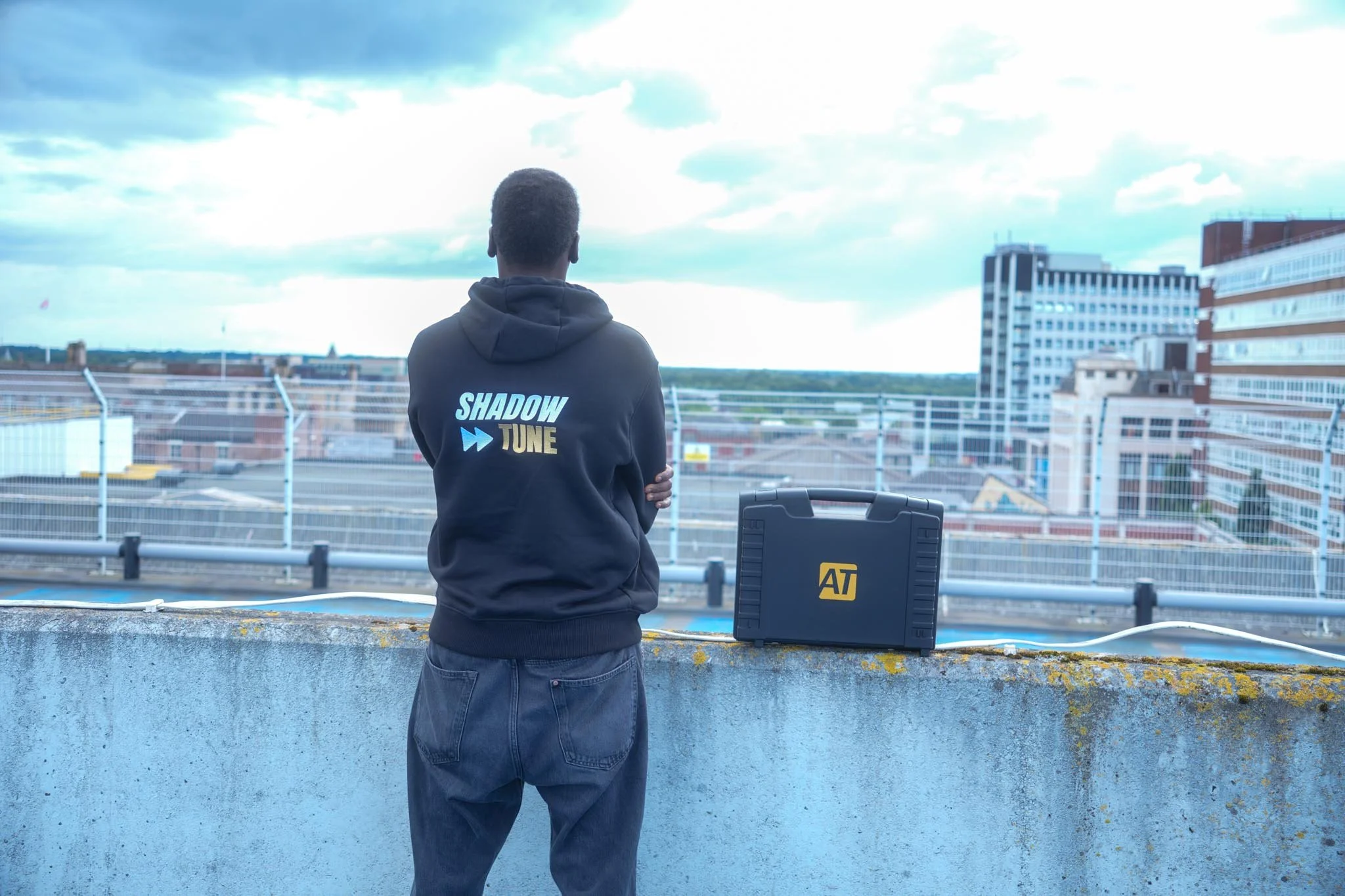 A person with short hair, wearing a black hoodie with "Shadow Tune" written on the back, stands on a rooftop or balcony, looking out over a cityscape with buildings and a cloudy sky.
