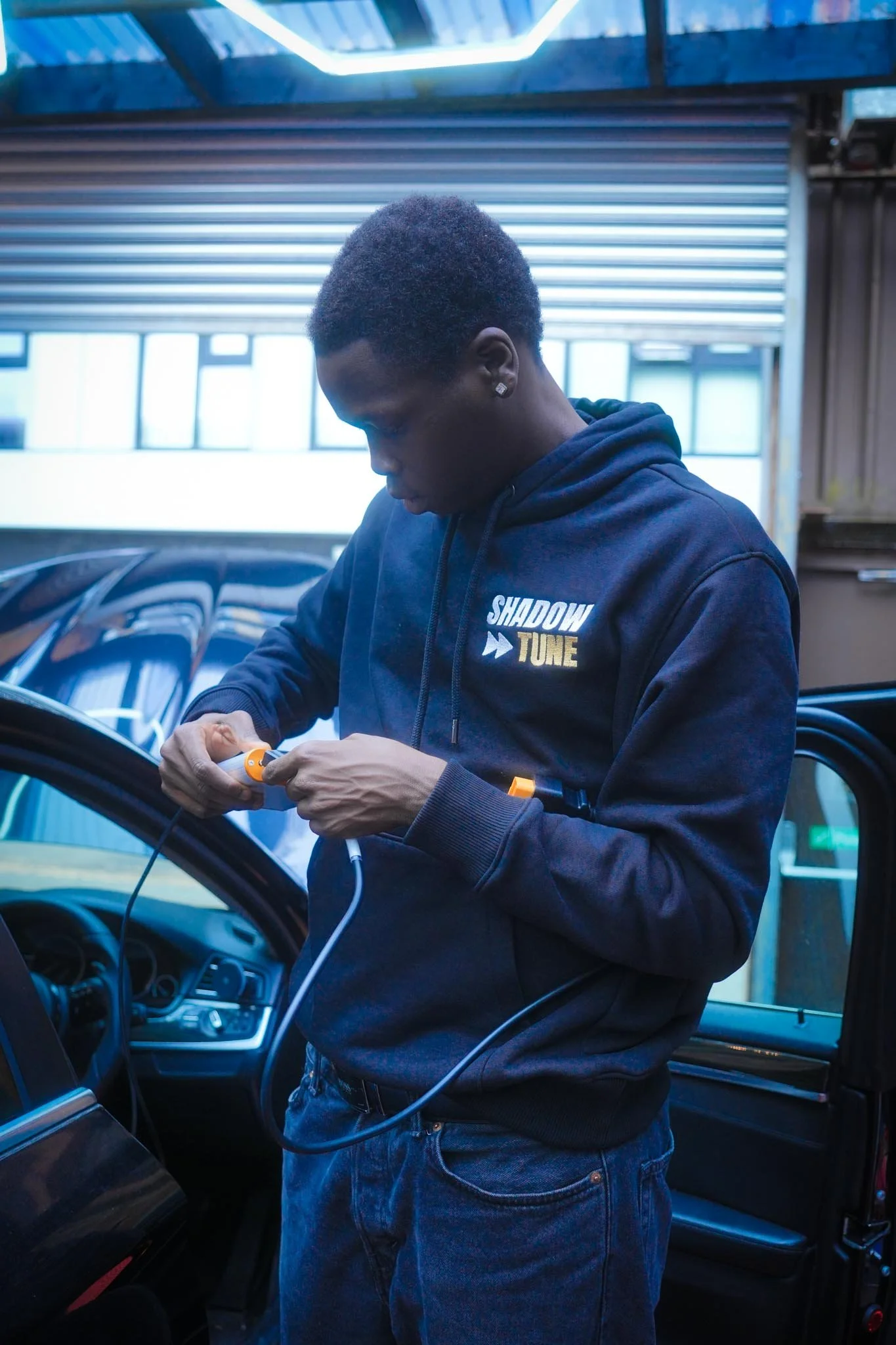 A young man with short curly hair and diamond earrings wearing a black hoodie with the words 'SHADOW TUNE' in white and yellow, standing inside a vehicle, working on a piece of equipment connected by a cable.
