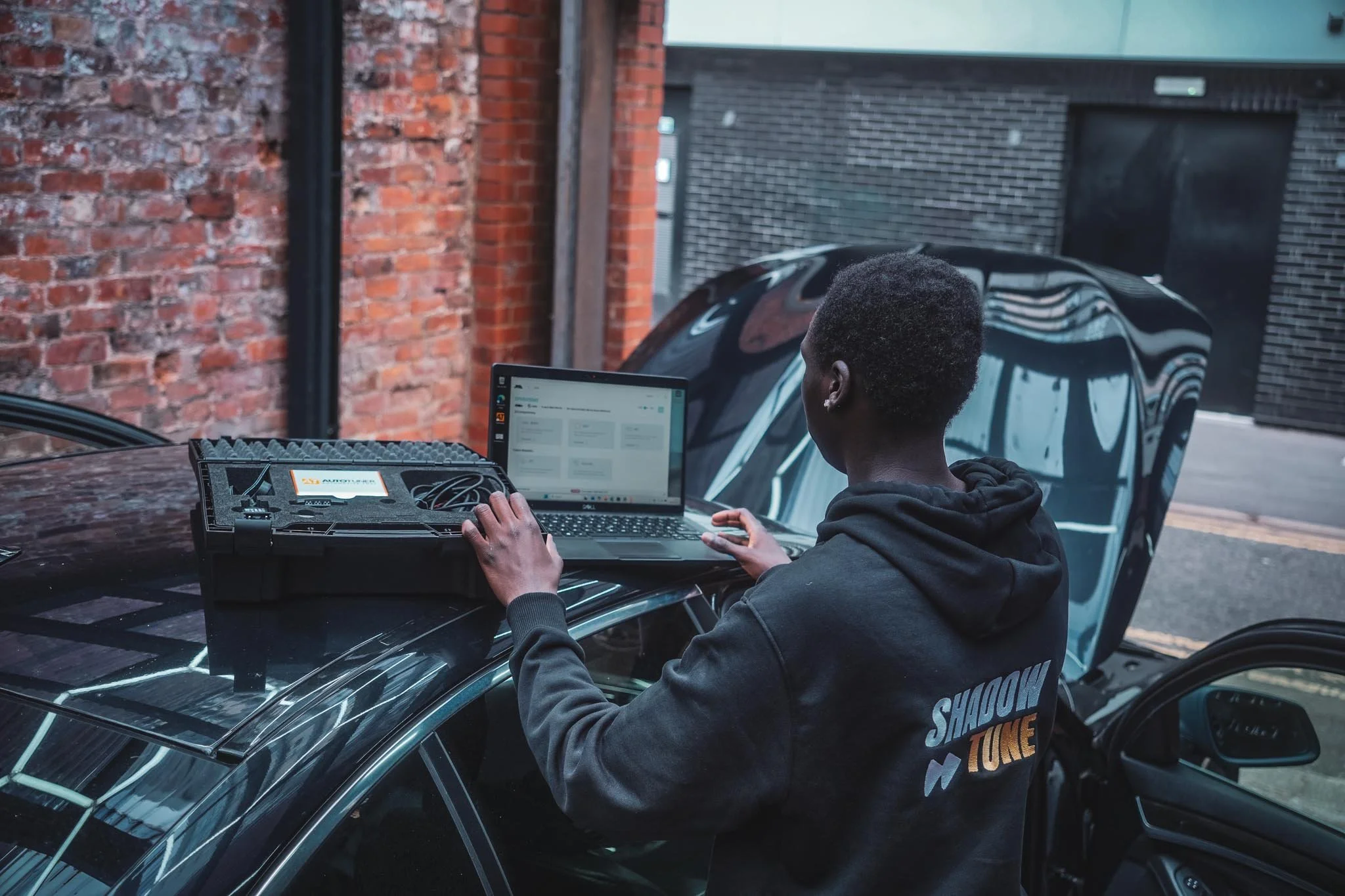 A person working on a laptop and electronic equipment at a table outdoors next to a brick building and a black sports car.