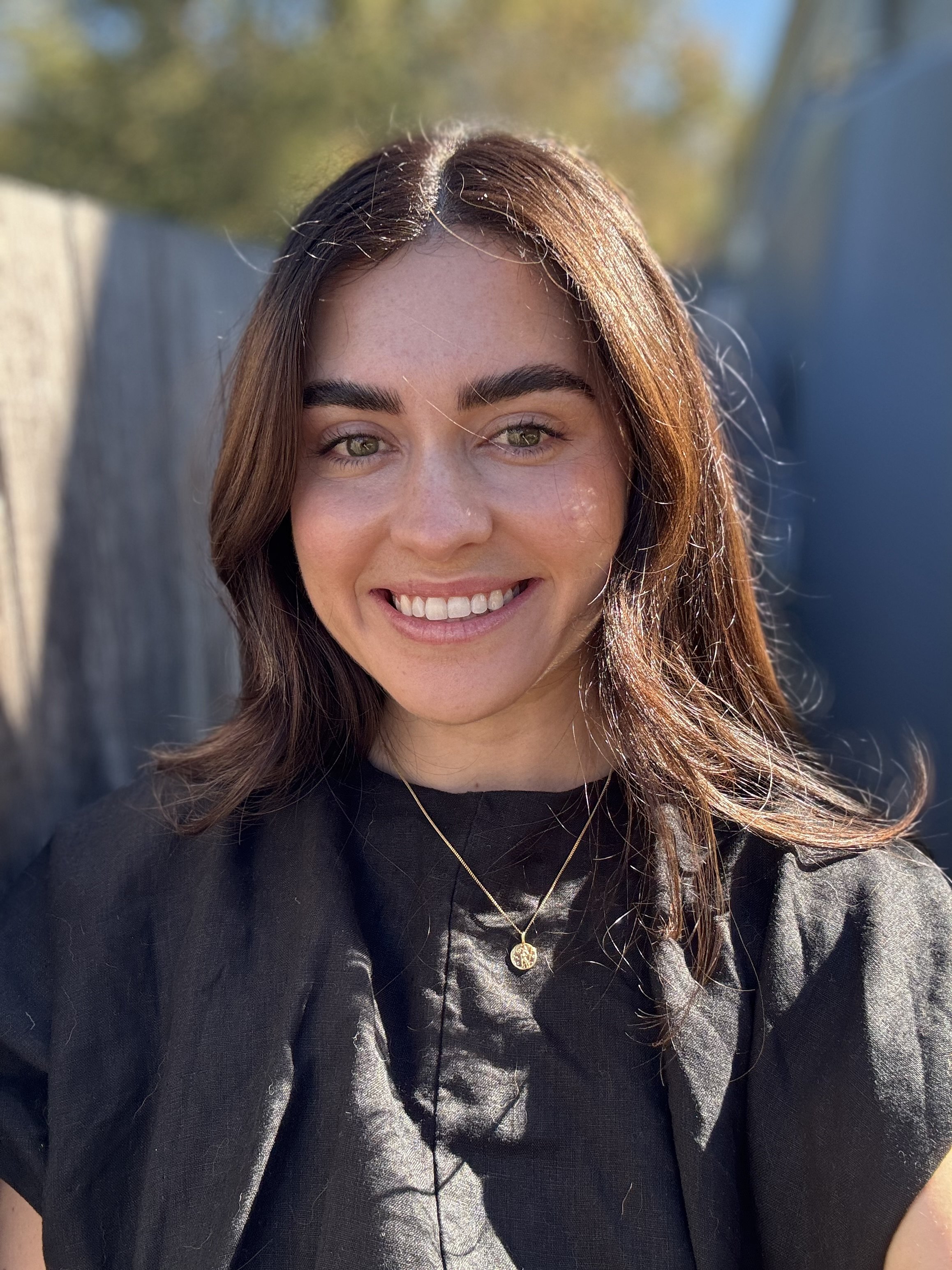 A young woman with long brown hair, green eyes, and a dark top smiling outdoors on a sunny day.