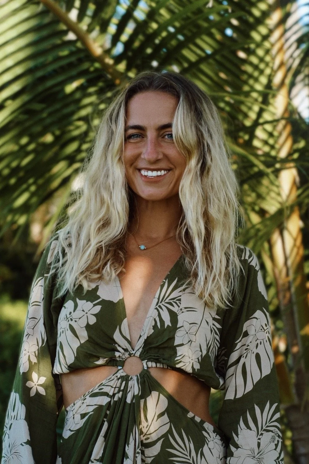 A smiling woman with long, wavy blonde hair stands outdoors surrounded by lush green tropical plants, wearing a green and white floral dress with a ring detail at the midsection.