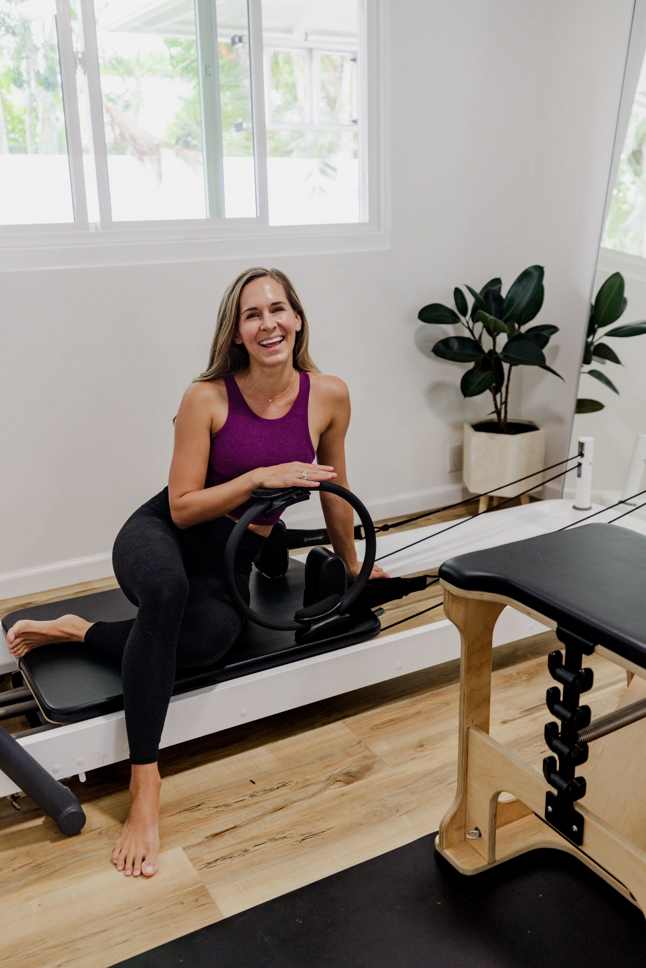 A woman in a purple sports bra and black leggings is kneeling on a Pilates reformer machine, smiling, with her left hand on the handle and her right hand resting on her thigh. The room has white walls, large windows with green foliage outside, and wooden flooring. There is a potted plant next to the woman and some exercise equipment nearby.