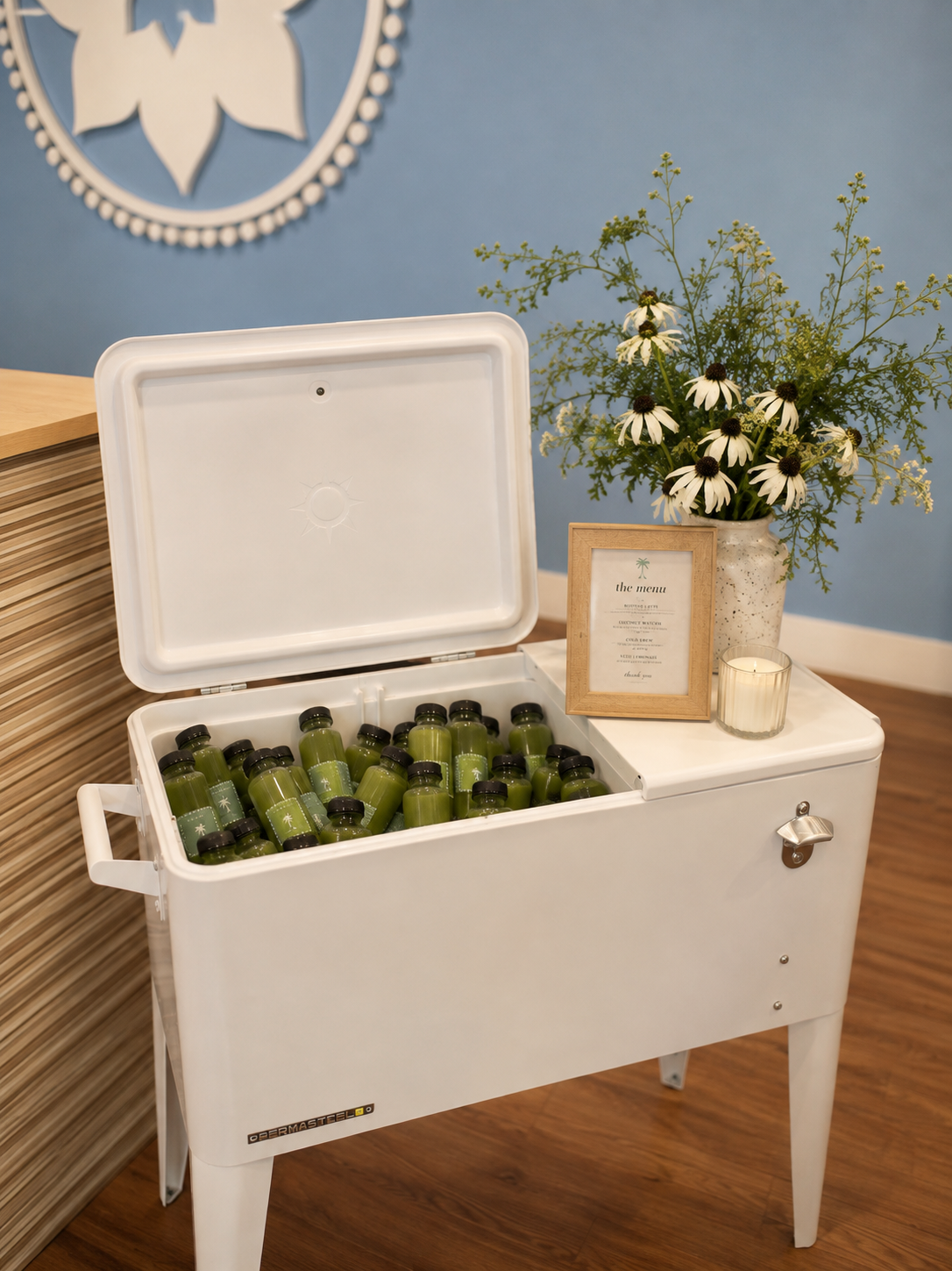 A white cooler filled with small green juice bottles, a framed menu, a vase with white daisies and green foliage, and a lit candle, set against a blue wall with a decorative emblem.