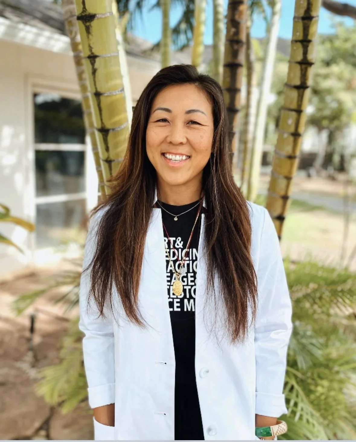 Woman with long dark hair, wearing a white lab coat and a black t-shirt with white text, smiling outdoors with green plants and trees in the background.