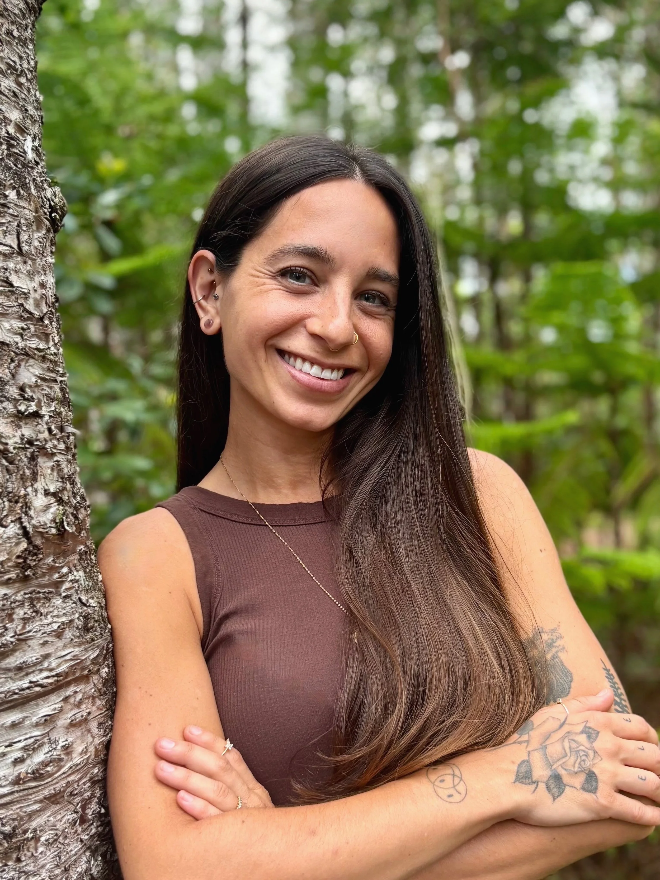 A smiling woman with long dark hair and tattoos on her arm, standing outdoors next to a tree in a green forest.