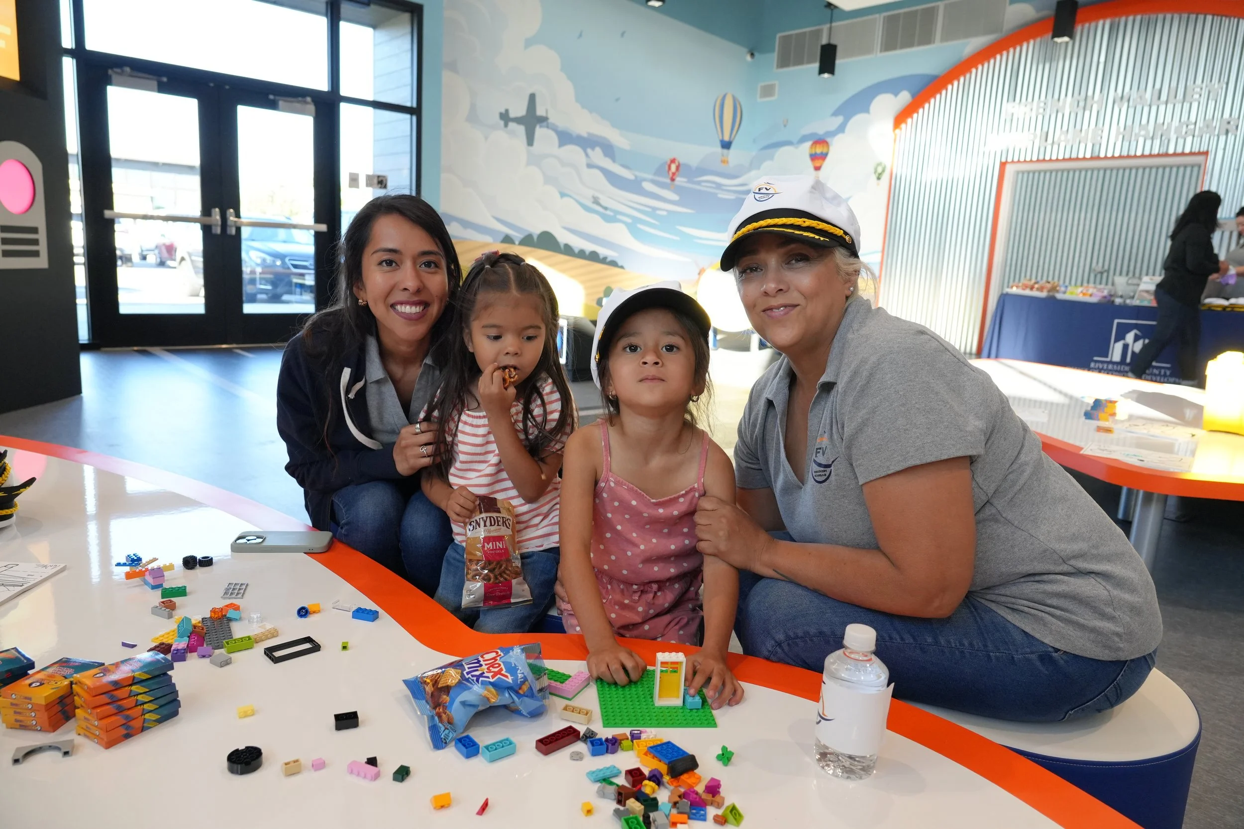 A family of four, including two young girls, is gathered around a table holding LEGO bricks, with a woman in a captain's hat and a man in a gray shirt. The background features a colorful mural with clouds, airplanes, and hot air balloons, indicating 