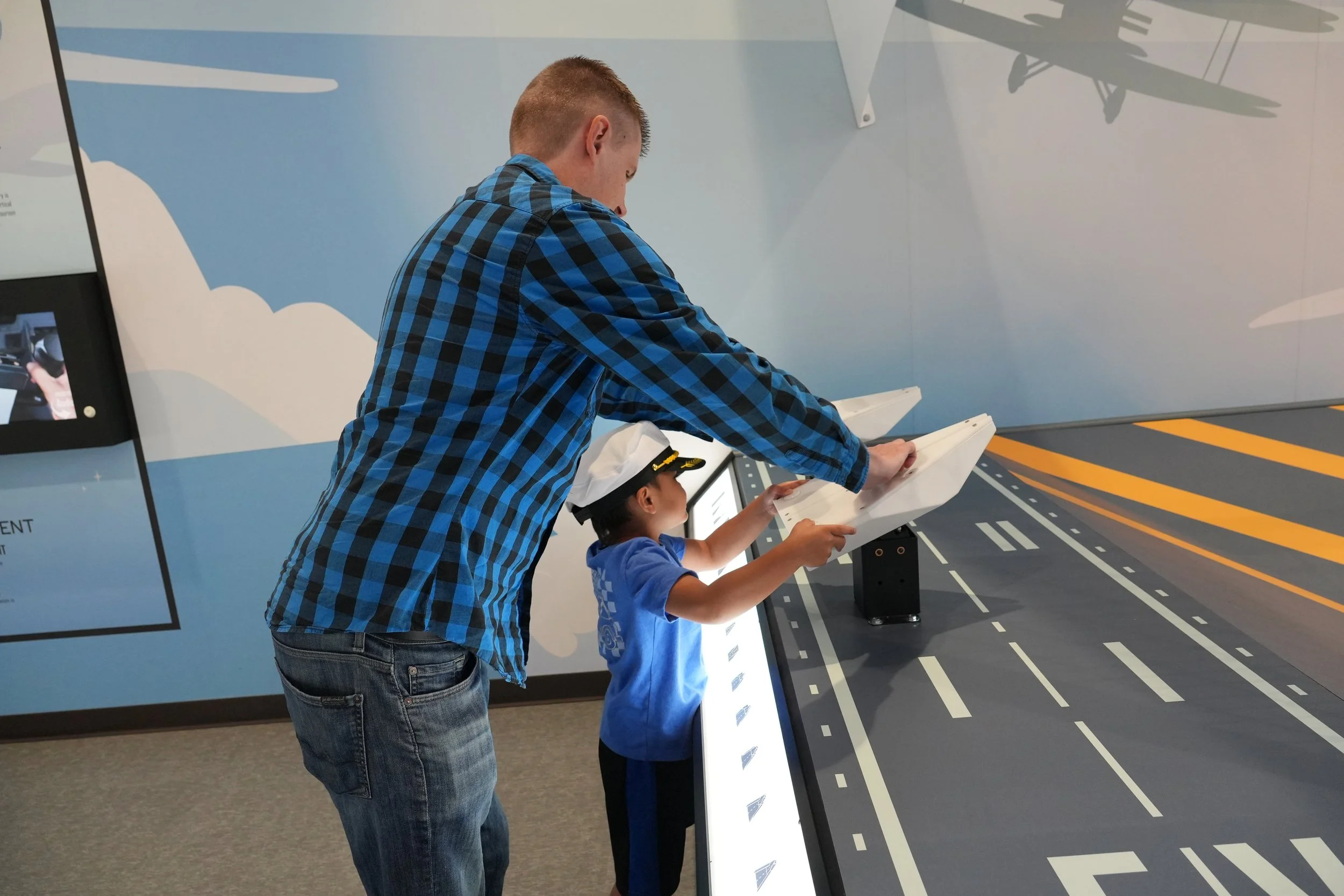 A man and a boy are interacting with an exhibit that simulates a road with lane markings. The man is leaning over the exhibit, and the boy is touching it while wearing a white hat.