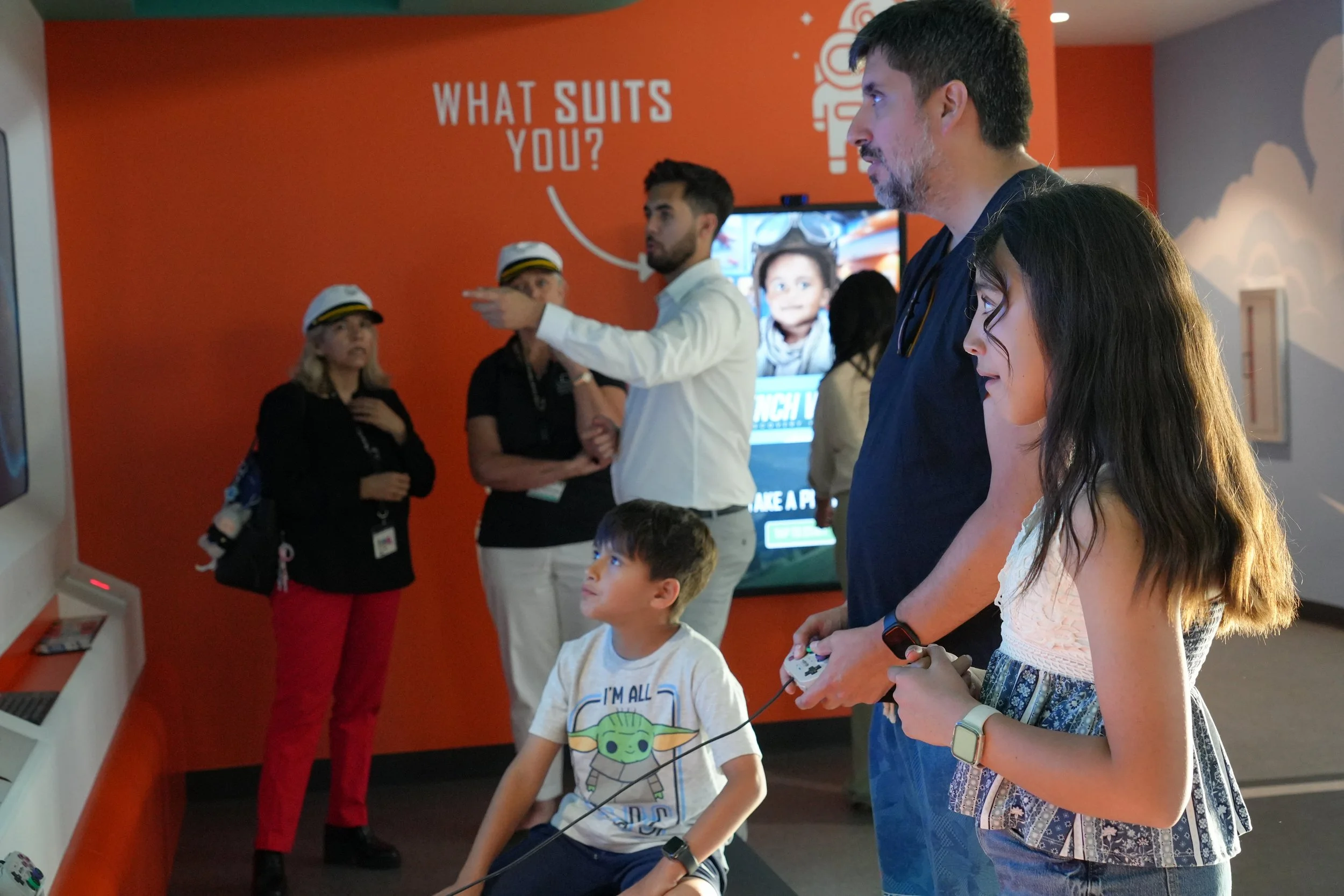 A group of people, including children and adults, are playing a video game in an entertainment area. There is a bright orange wall with the text 'What suits you?' in the background. One man is holding a game controller, and others are watching or waiting their turn.