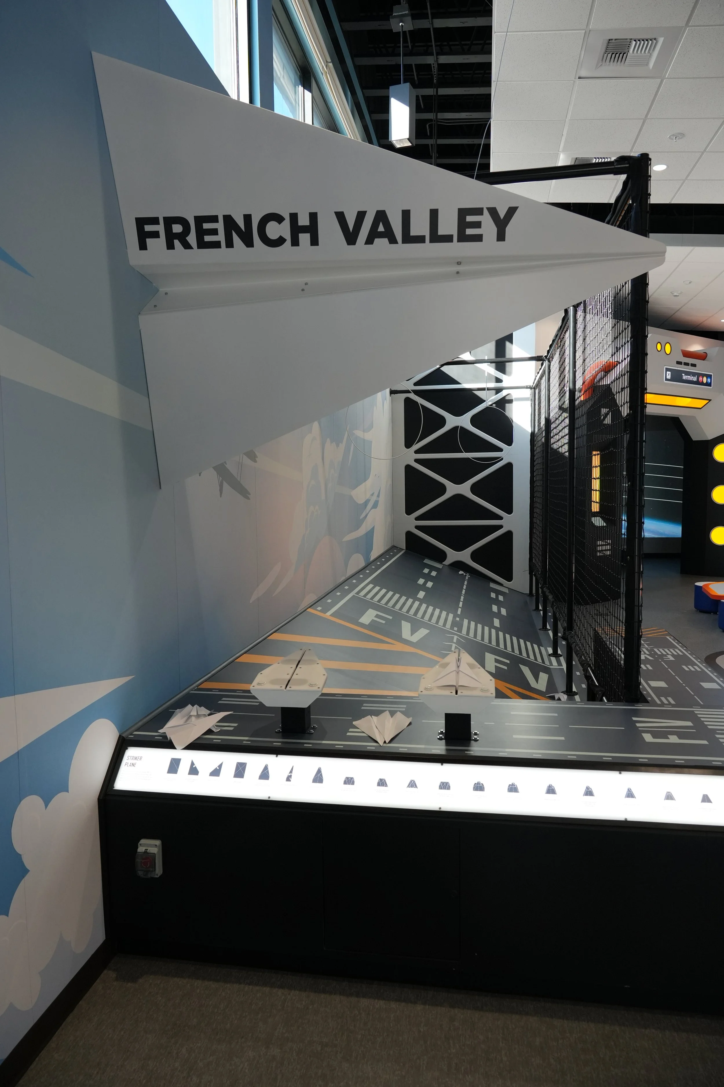 Indoor area with a white sign reading 'FRENCH VALLEY' hanging from the ceiling, black safety barrier fencing, a display with paper airplanes, and a floor graphic resembling an airport runway.