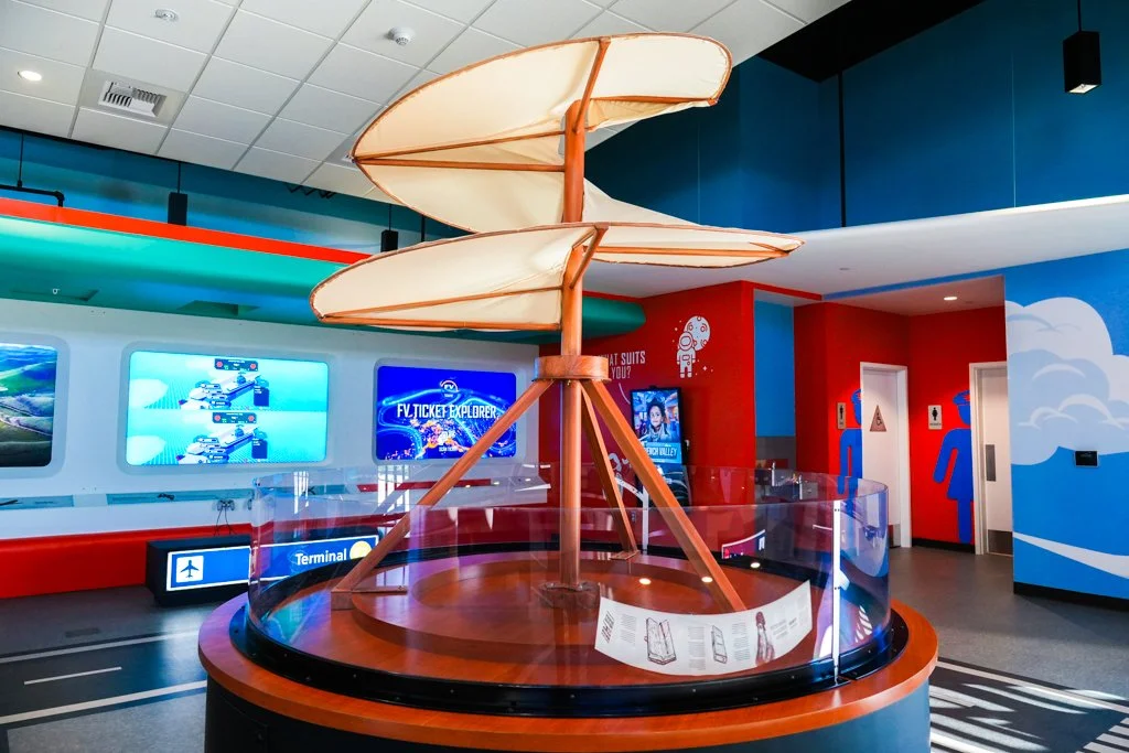 Indoor playground featuring a large spiral slide with beige fabric and orange supports, surrounded by a clear safety barrier, in a colorful children's entertainment area with digital screens and bathroom signs.