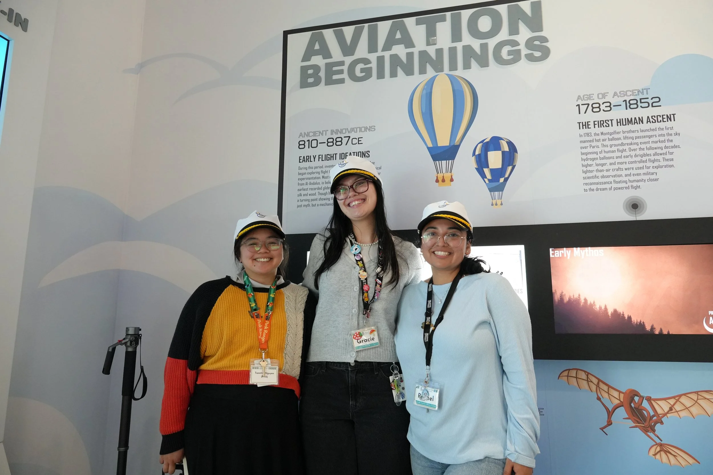 Three women wearing glasses and badges, standing together at an aviation exhibition. Behind them is a wall with information about the history of flight, including images of hot air balloons and a dinosaur illustration.