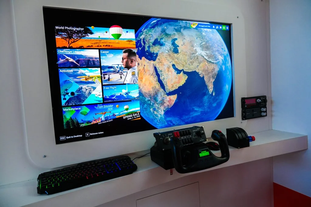 Large monitor displaying a world map and a video game menu with flight-themed options, a keyboard, a joystick, and a racing wheel controller on a white desk, with a white wall in the background.