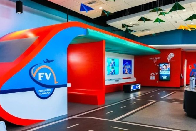 Indoor play area with a colorful airplane-themed structure, digital screens, and playful decorations including hanging green and blue shapes.