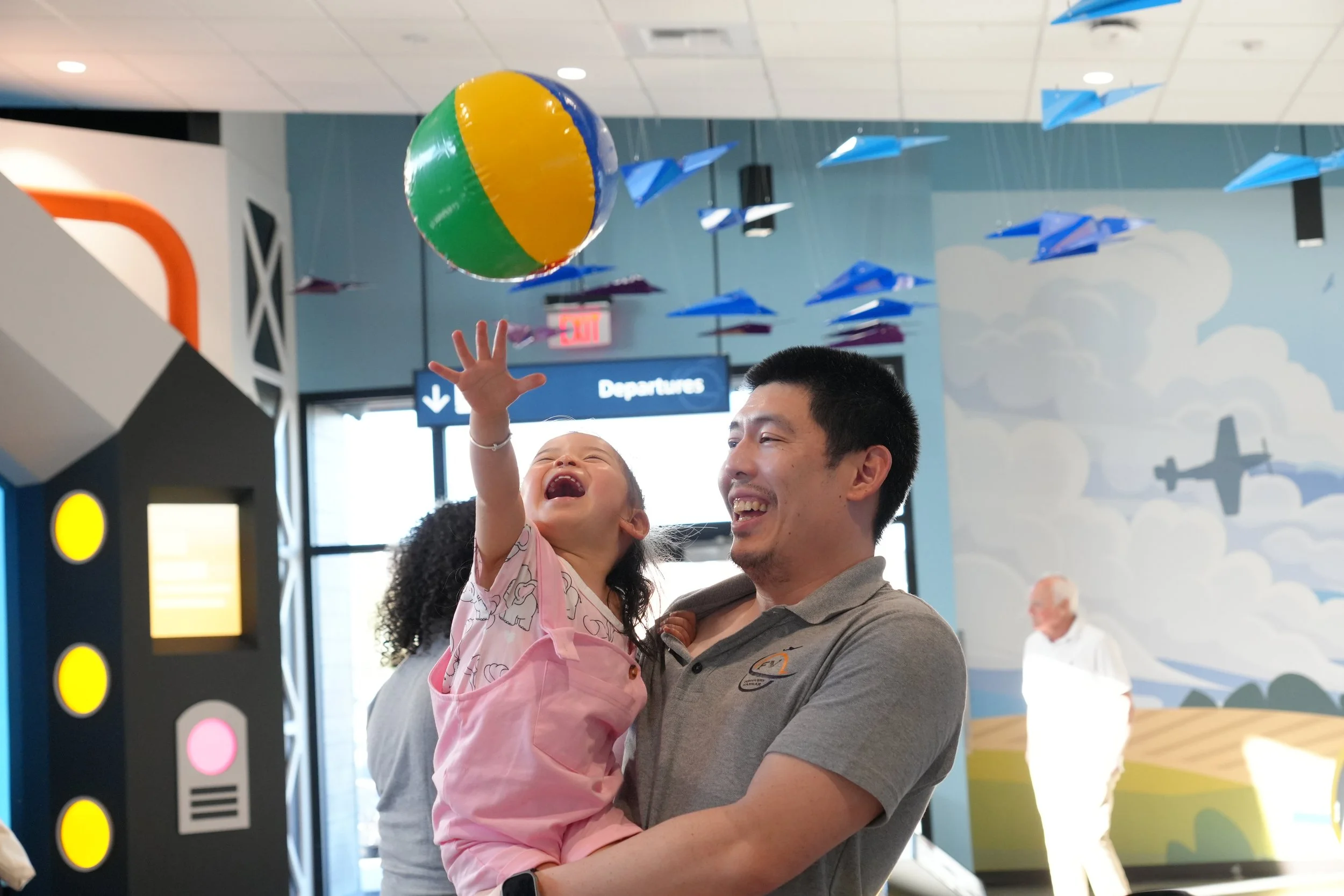 A man holding a young girl at an indoor airport or transportation hub, with colorful paper airplanes and a beach ball overhead, and a departures sign in the background.