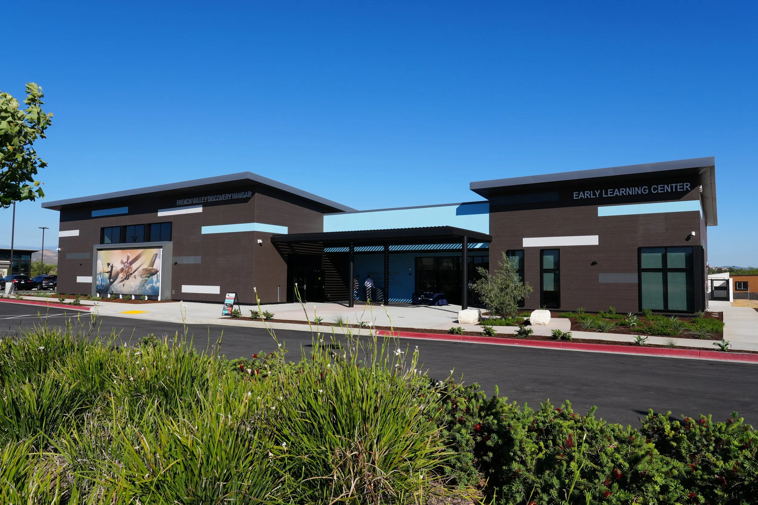 Modern school building with signs for French Valley Discovery Hangar and Early Learning Center, surrounded by landscaped greenery under a clear blue sky.