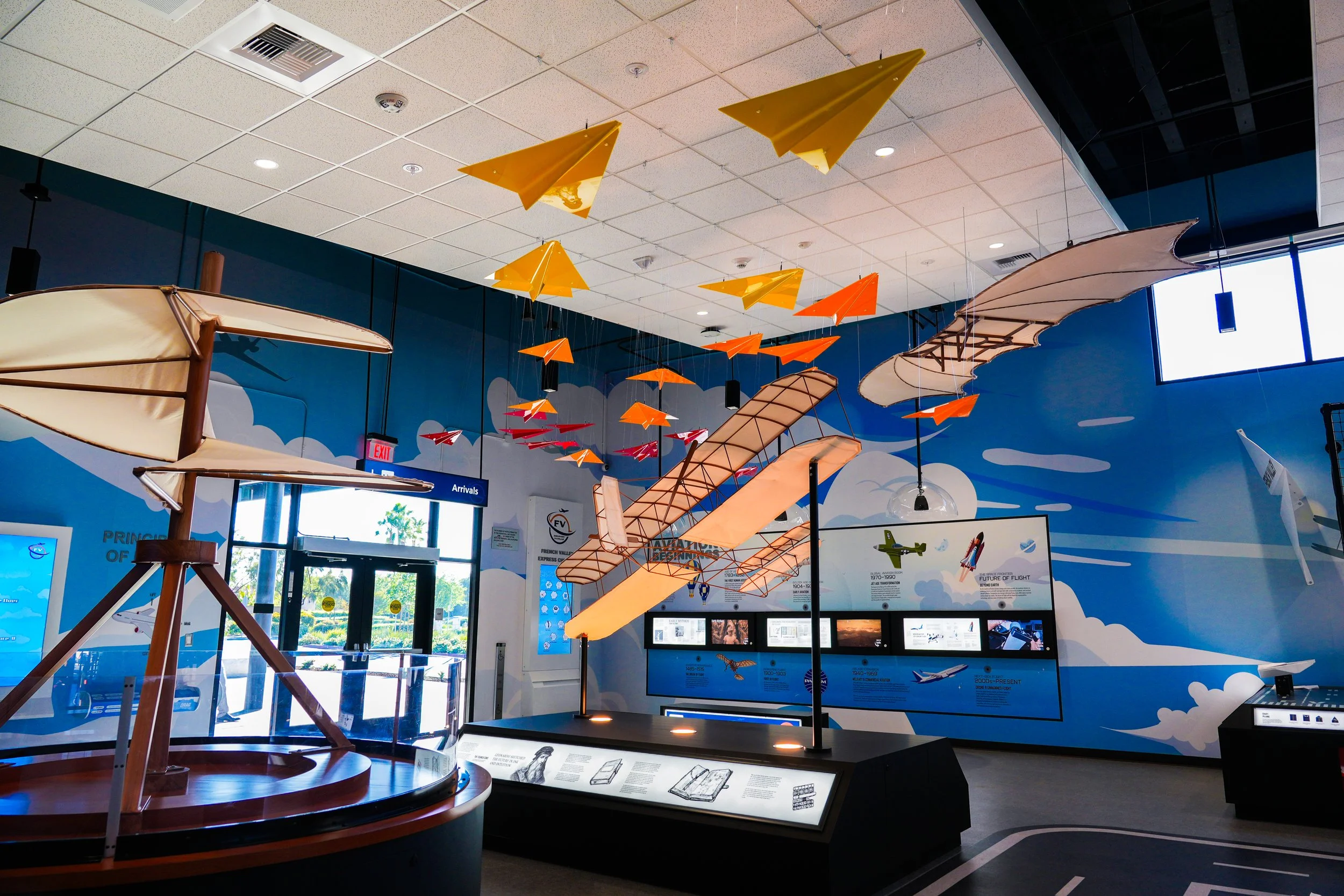 Indoor exhibit with suspended model airplanes hanging from the ceiling, colorful and in various sizes, in a space with blue walls and informational displays.