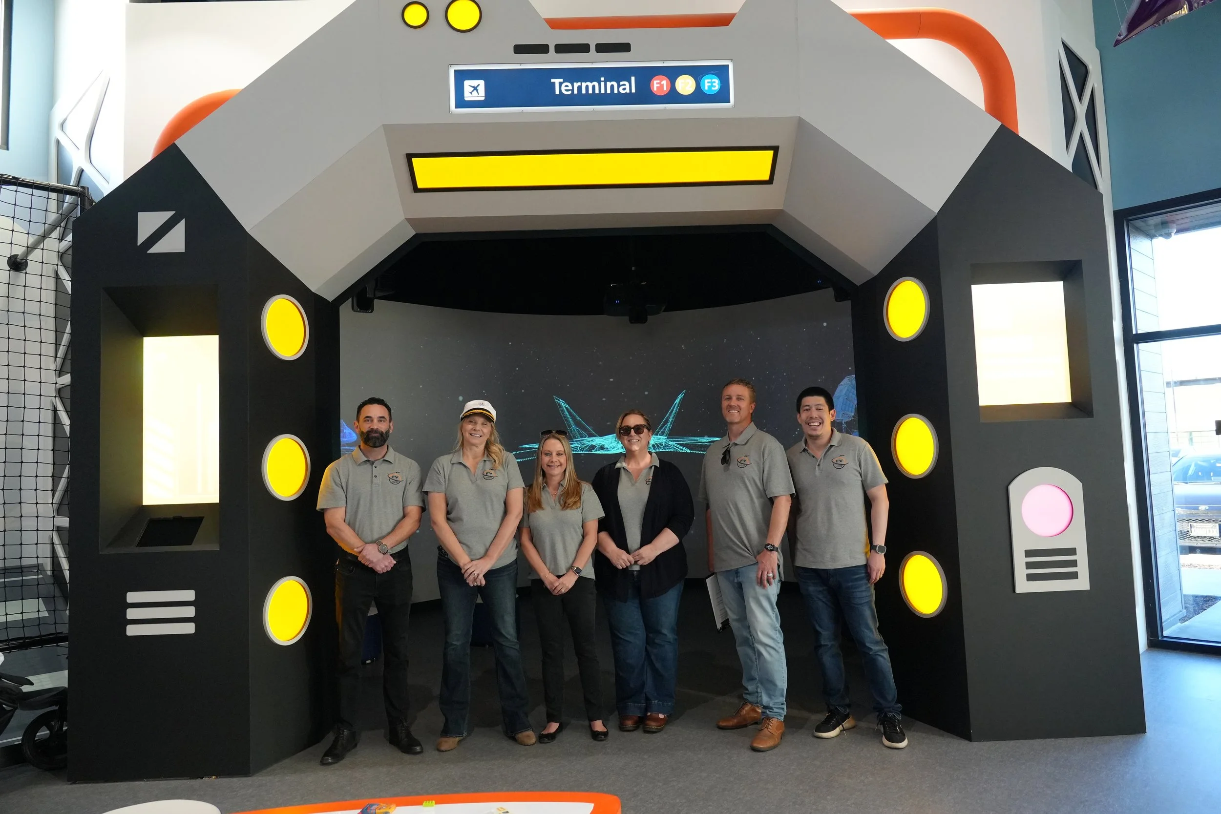 A group of six people standing in front of a space-themed photo booth at an airport terminal. The booth has a black and white design with yellow lights, and a sign labeled 'Terminal' with airline codes. The group appears to be smiling and posing for 