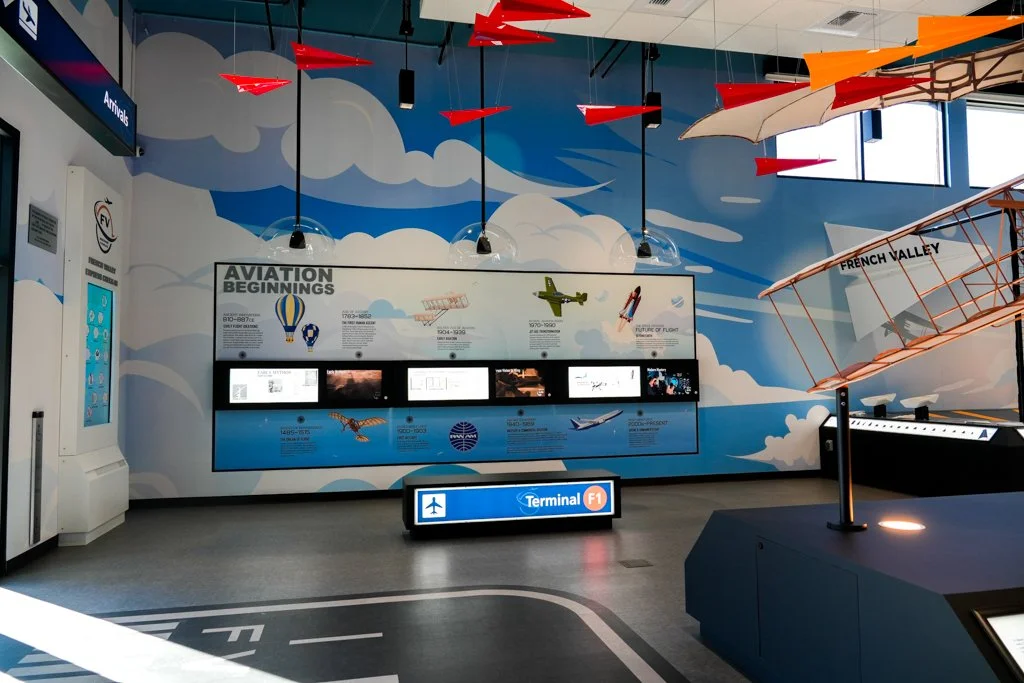 Indoor aviation exhibit with airplane models and a large wall display titled 'Aviation Beginnings' at an airport terminal.