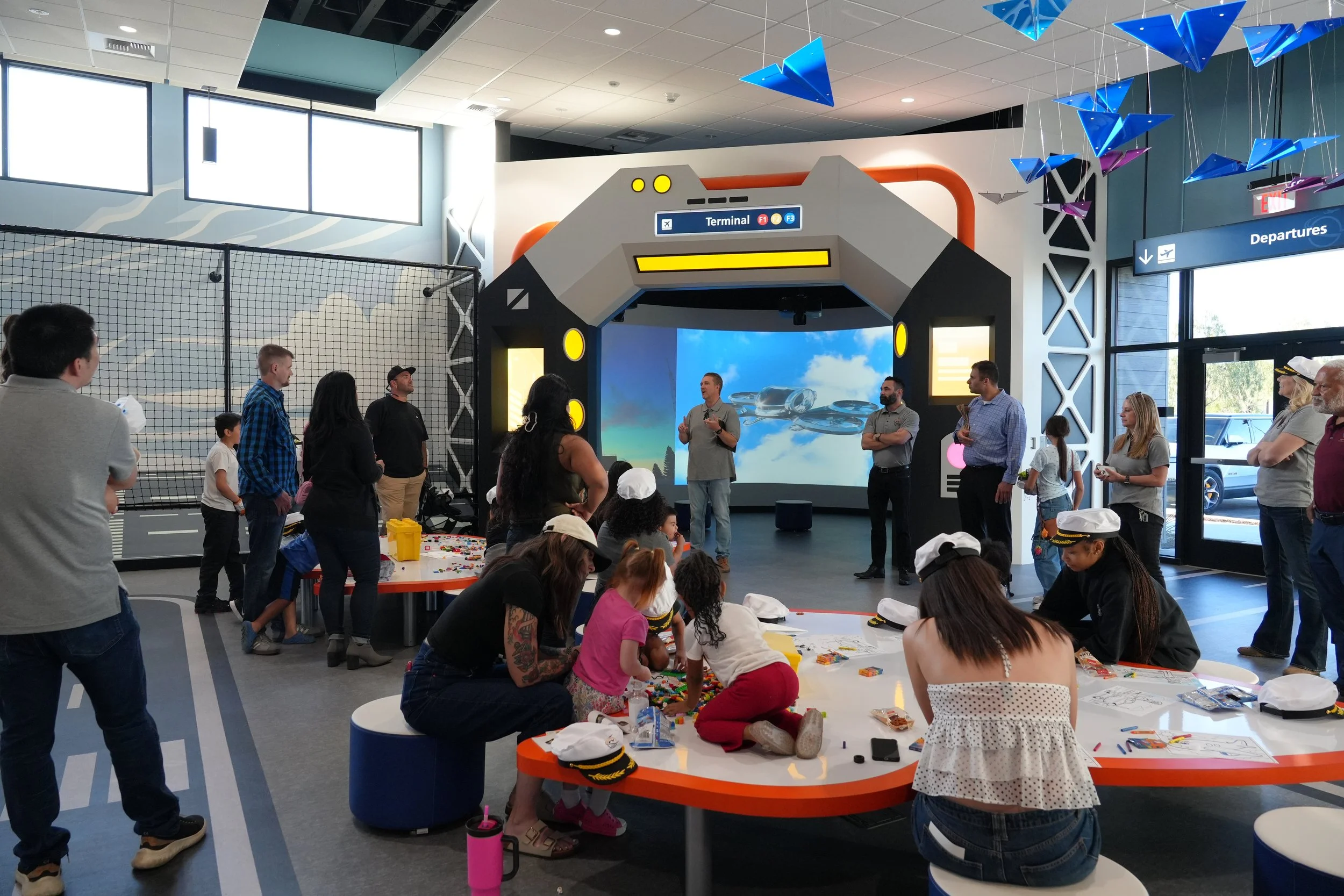 Children and adults at a science or technology event inside an airport-themed area with a large spaceship-shaped installation labeled 'Terminal' and a Departure sign. People are engaging in activities, some wearing hats resembling pilot caps.