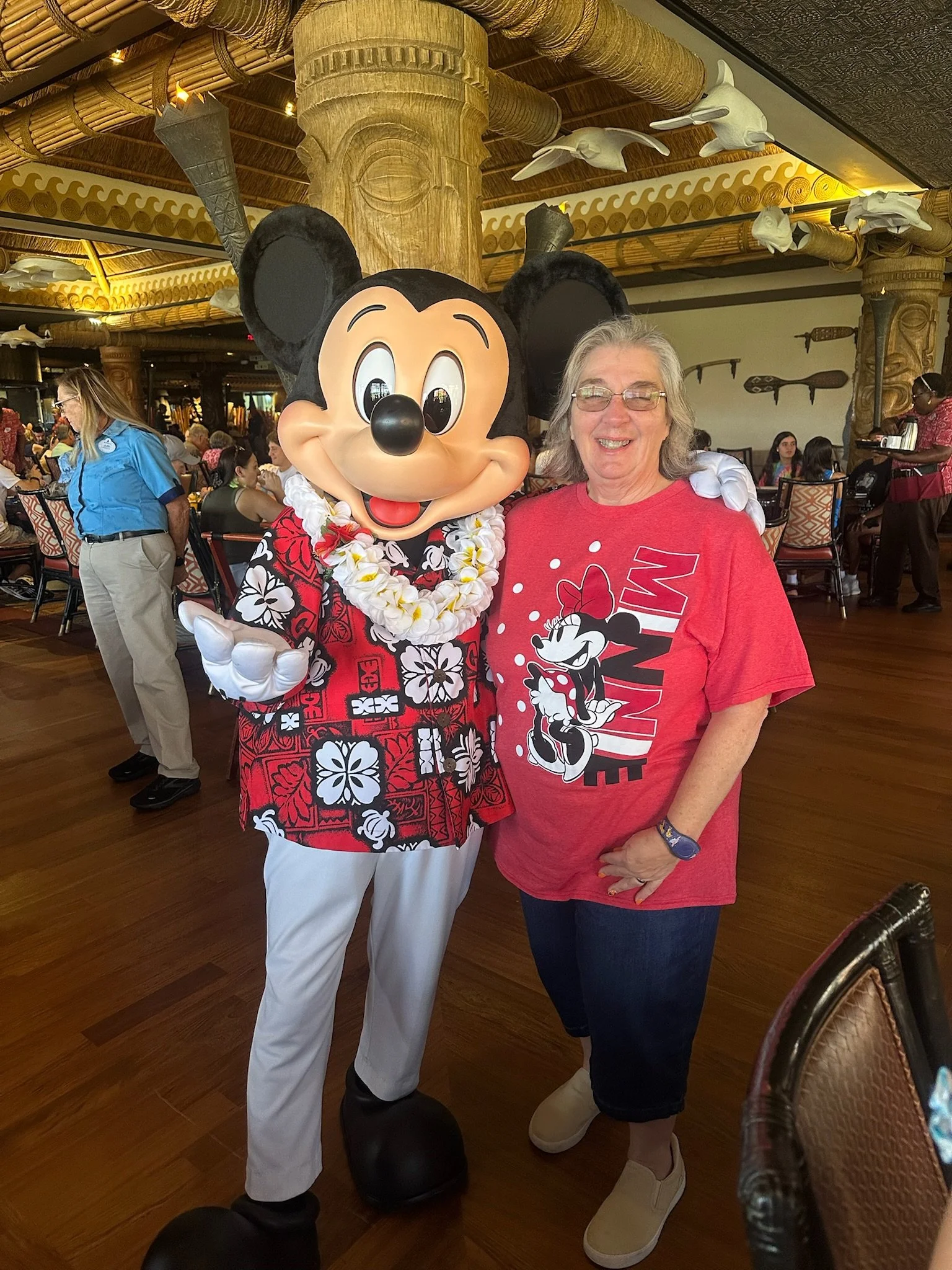 A woman wearing a red Minnie Mouse t-shirt standing next to a person dressed as Mickey Mouse in a Hawaiian shirt and lei at a Disney theme park restaurant. The background features wooden decor and people dining.