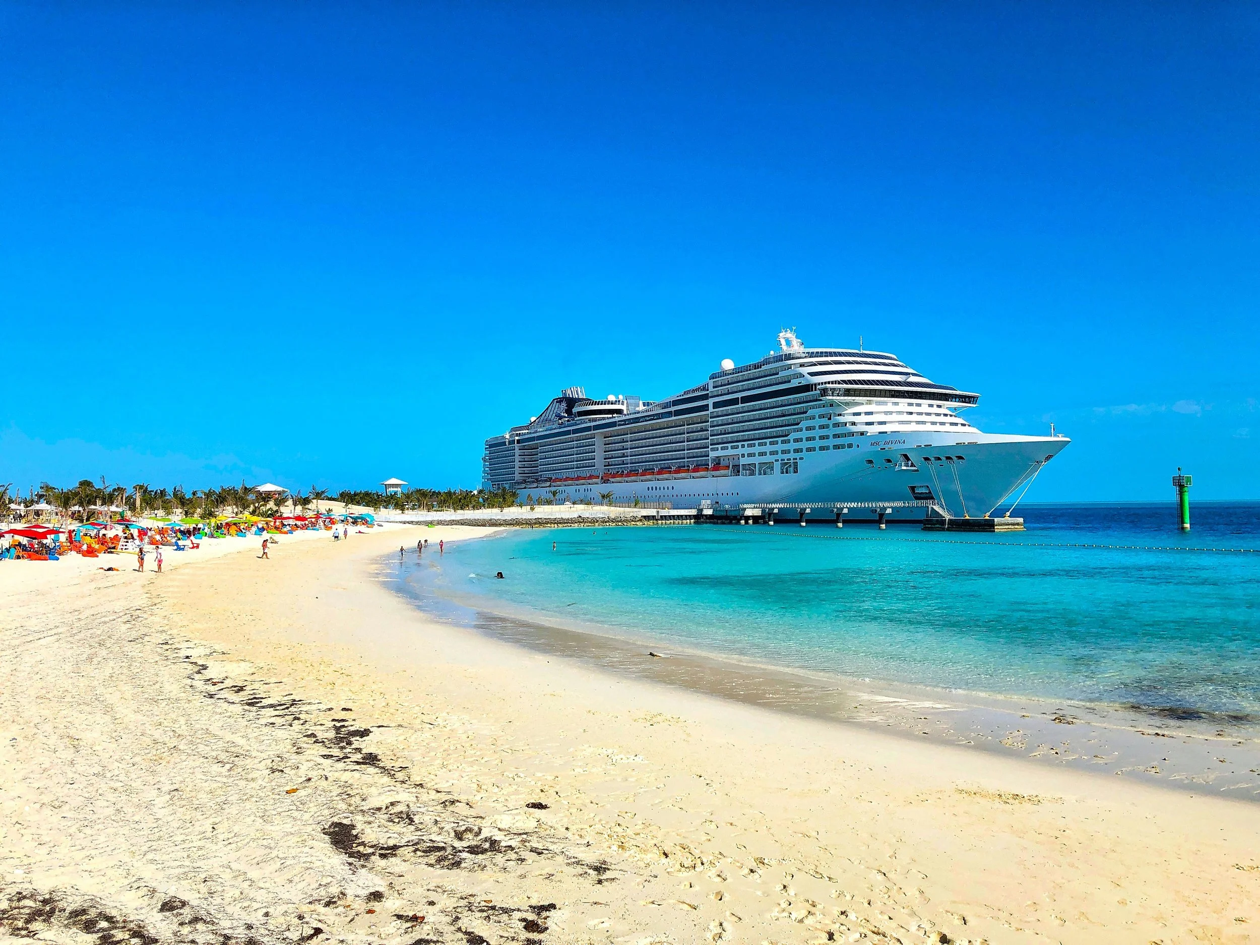 A large cruise ship docked at a tropical beach with white sand, colorful umbrellas, and people relaxing and swimming in the clear blue water, under a bright blue sky.