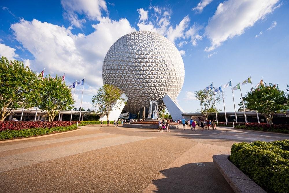 Epcot ball on a sunny day with a few clouds and visitors walking nearby.