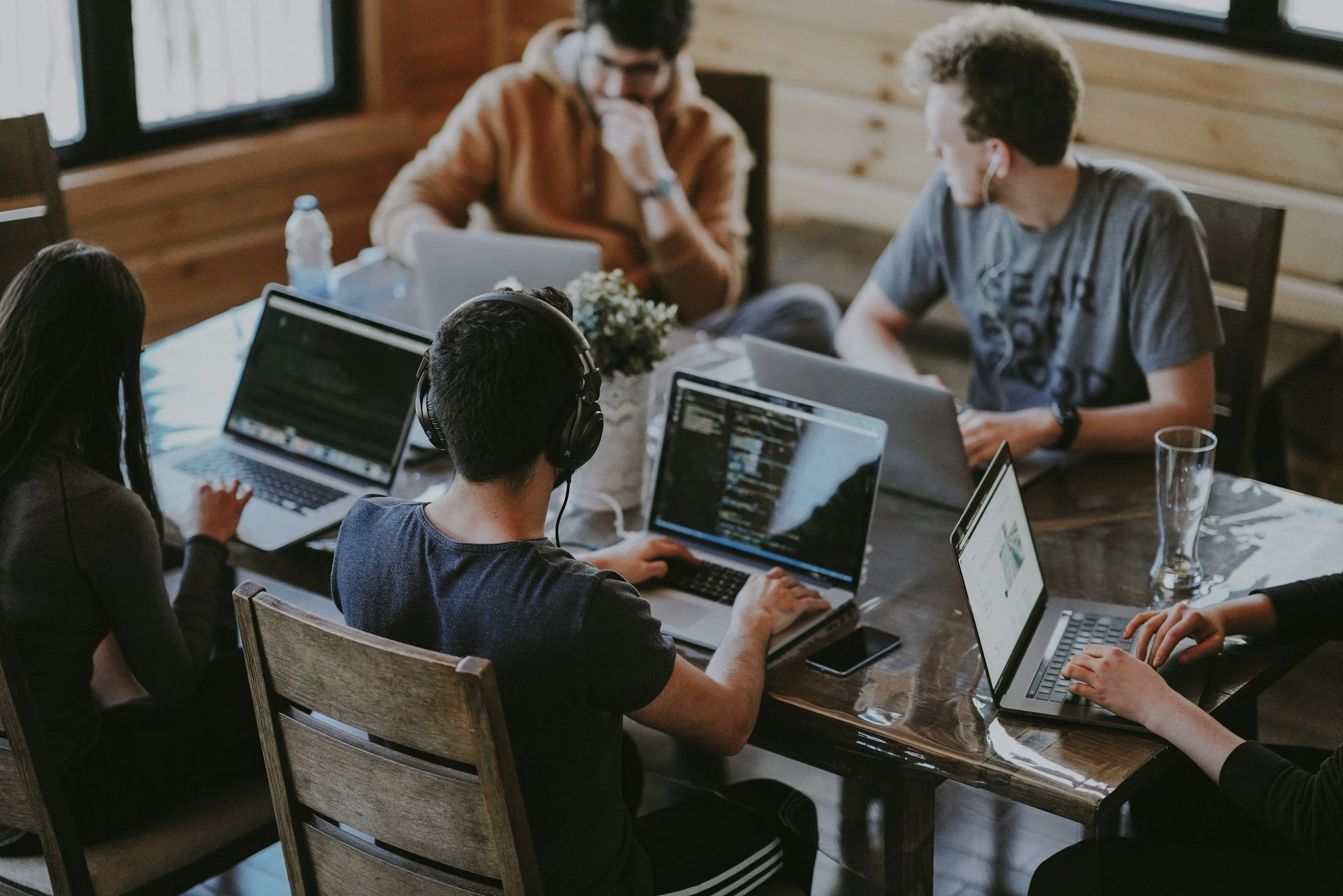 A group of young people working together at a wooden table with laptops, one person wearing headphones, in a cozy wooden-paneled room.
