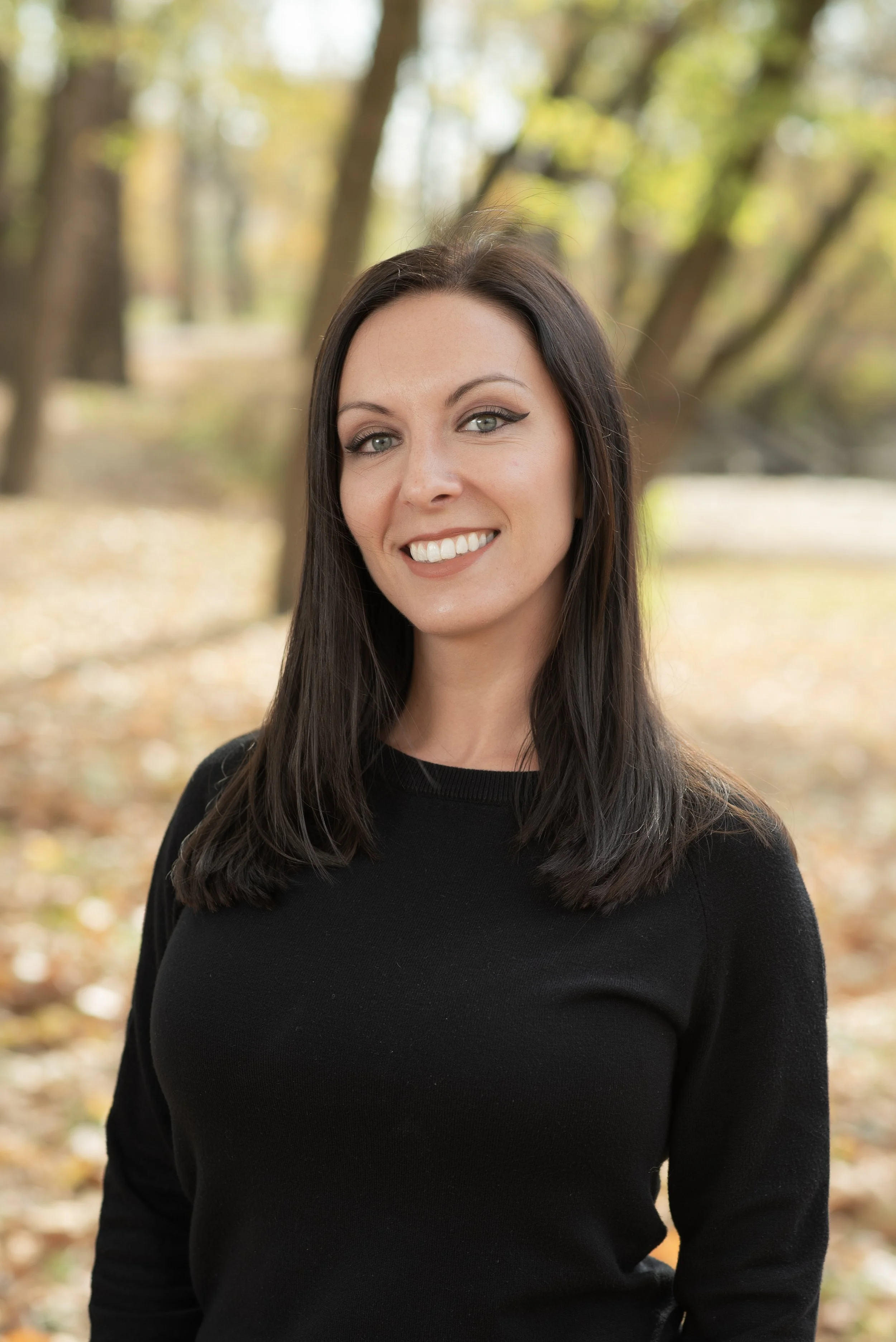 A woman with long dark hair wearing a black top, smiling, outdoors in a park with autumn leaves and trees in the background.