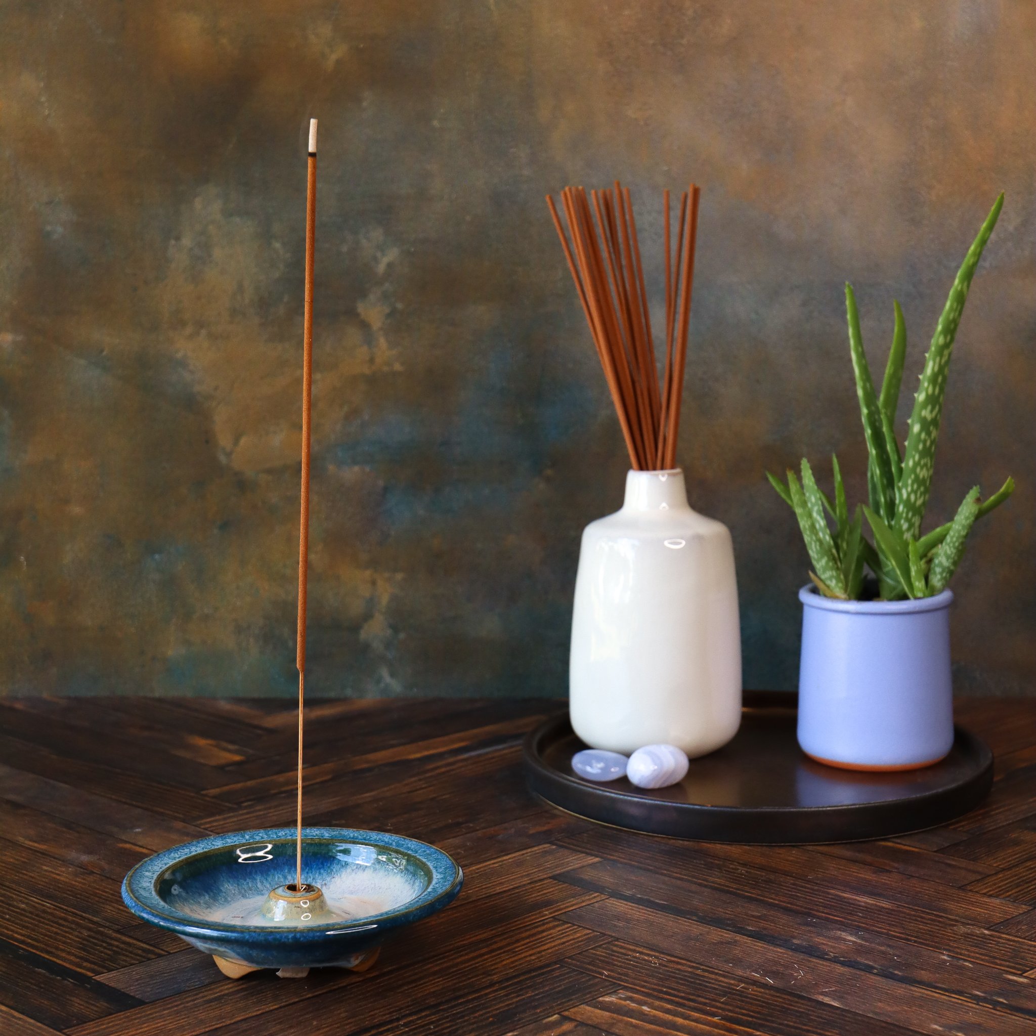 A brown incense stick in a small blue ceramic dish, a bundle of thin brown incense sticks in a white vase, and a potted aloe vera plant on a dark wooden table against a textured brown wall.
