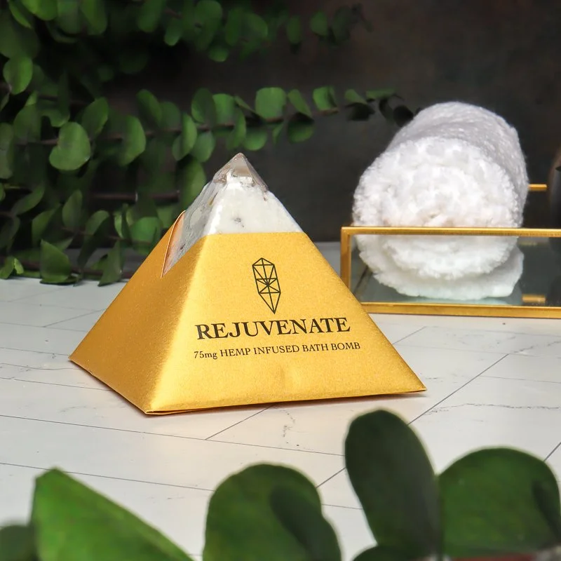 A pyramid-shaped bath bomb in gold packaging labeled 'REJUVENATE, 75mg Hemp Infused Bath Bomb' with a white fluffy towel rolled on a small glass tray in the background, and greenery in the setting.