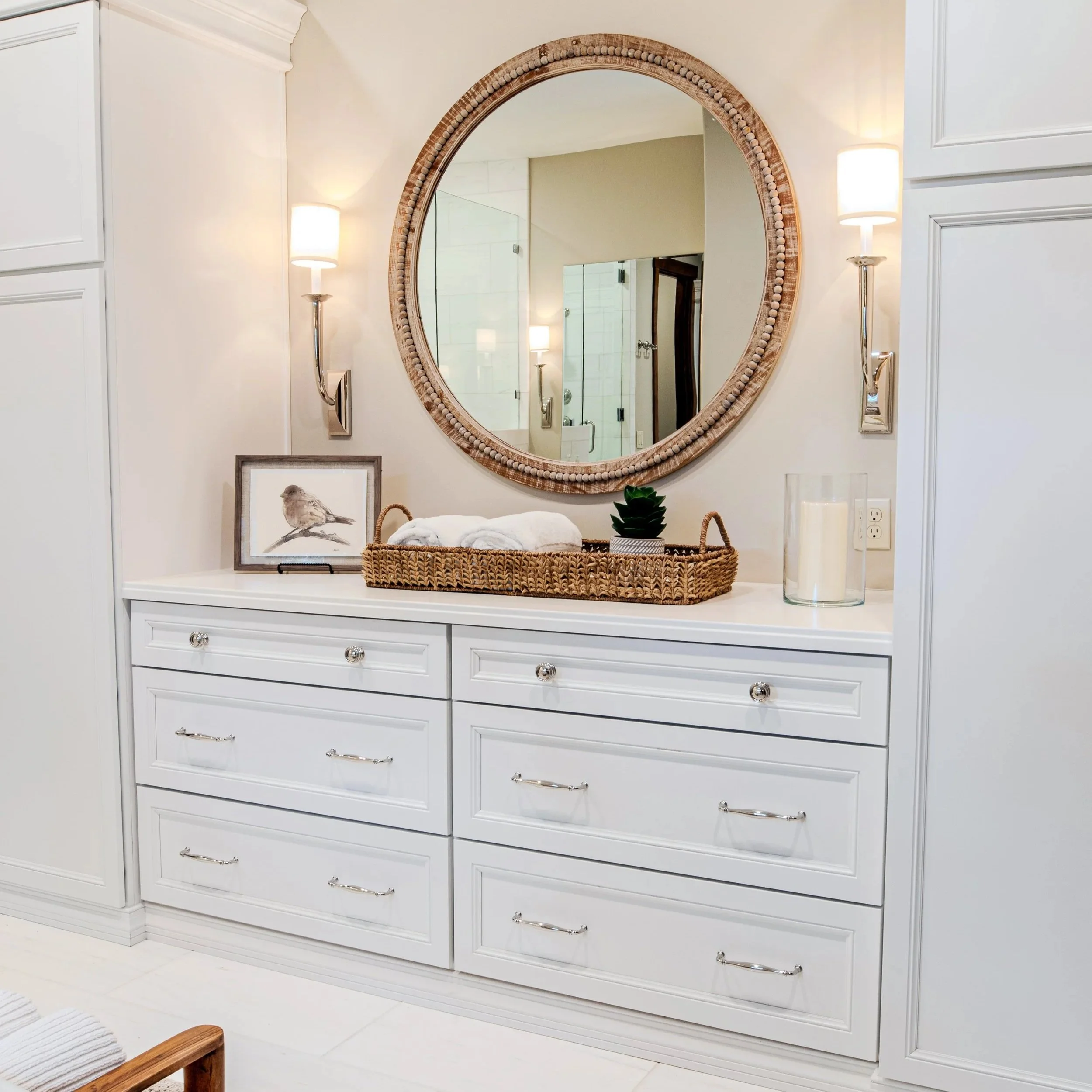 A round mirror with beads around the frame mounted on a wall above a white dresser. The dresser has drawers with shiny handles, a framed bird illustration, a woven basket, a small plant, and two candles on top. The room has white walls, a white ceiling, and white floor tiles.