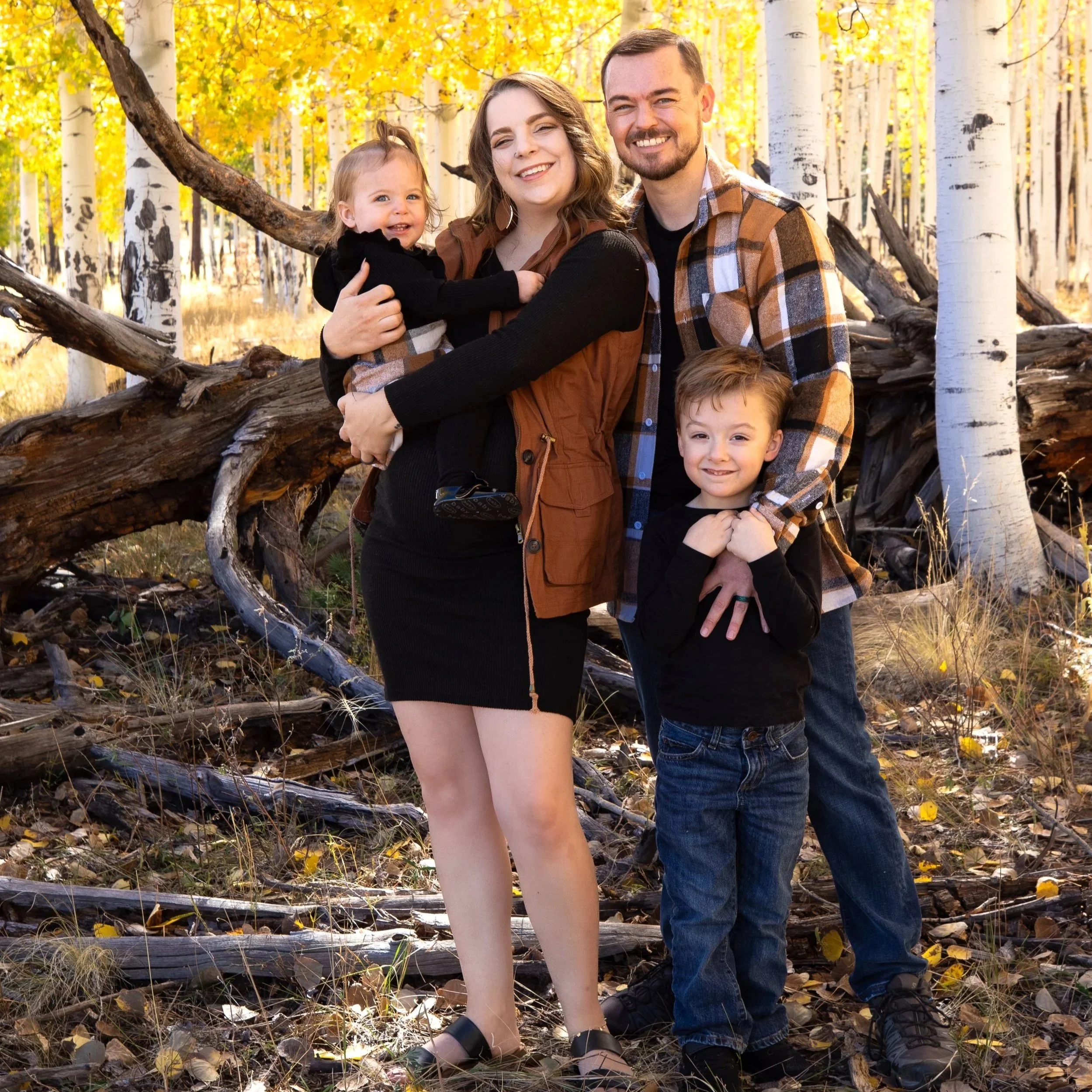 A family of four enjoying a fall day in a forest with golden trees. The mother, father, son, and one toddler girl are smiling and posing for a photo. The mother has shoulder-length brown hair, the father has short brown hair and a beard, the boy has short light brown hair, and the girl has blonde hair tied up. They are dressed in casual fall clothing, and the scene is filled with fallen yellow leaves and white birch trees in the background.