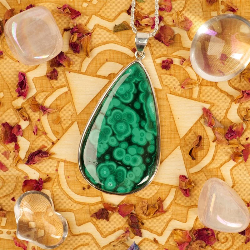 A teardrop-shaped green malachite pendant necklace surrounded by scattered dried flower petals and clear glass gemstones on a wooden patterned surface.