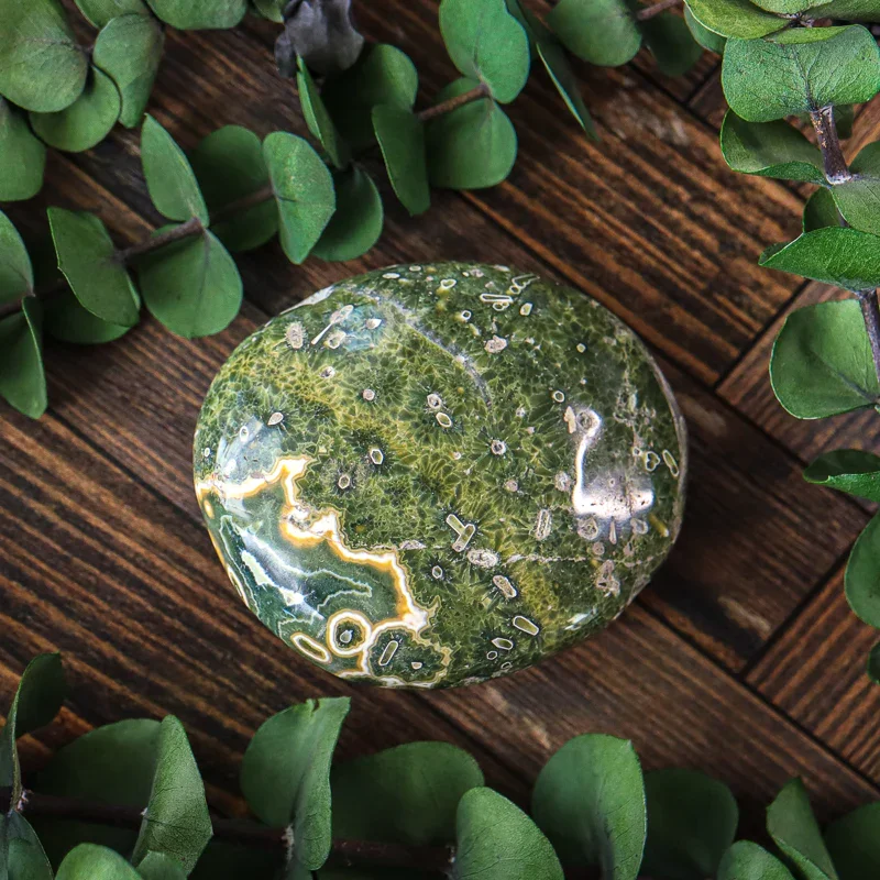 A polished green and yellow ornamental stone with intricate patterns, placed on a wooden surface surrounded by green foliage.