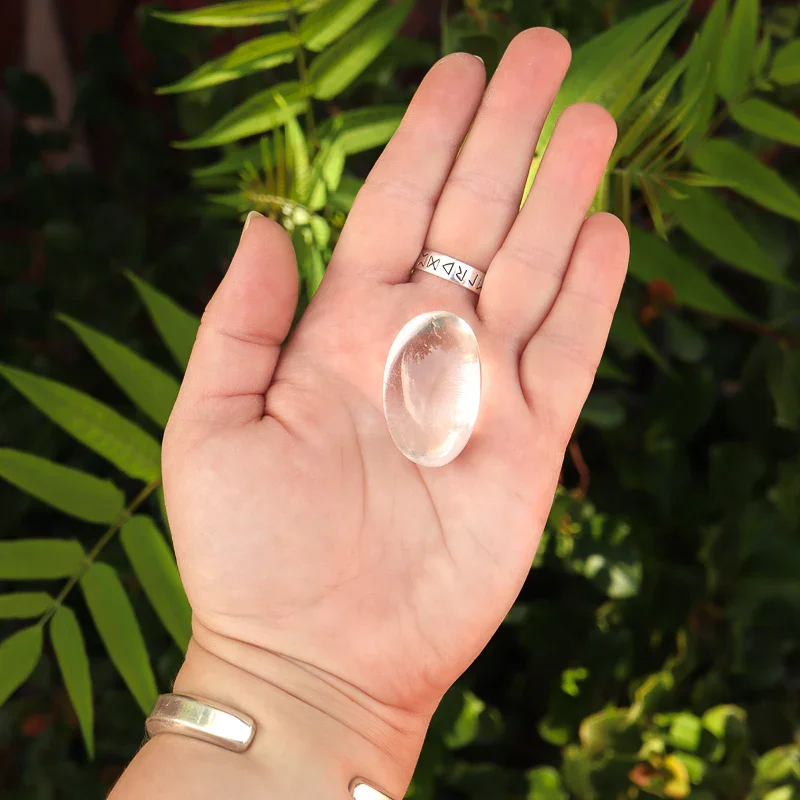 Hand holding a large, oval-shaped, clear glass or crystal gem against a background of green leaves, wearing rings on the fingers and wrist.