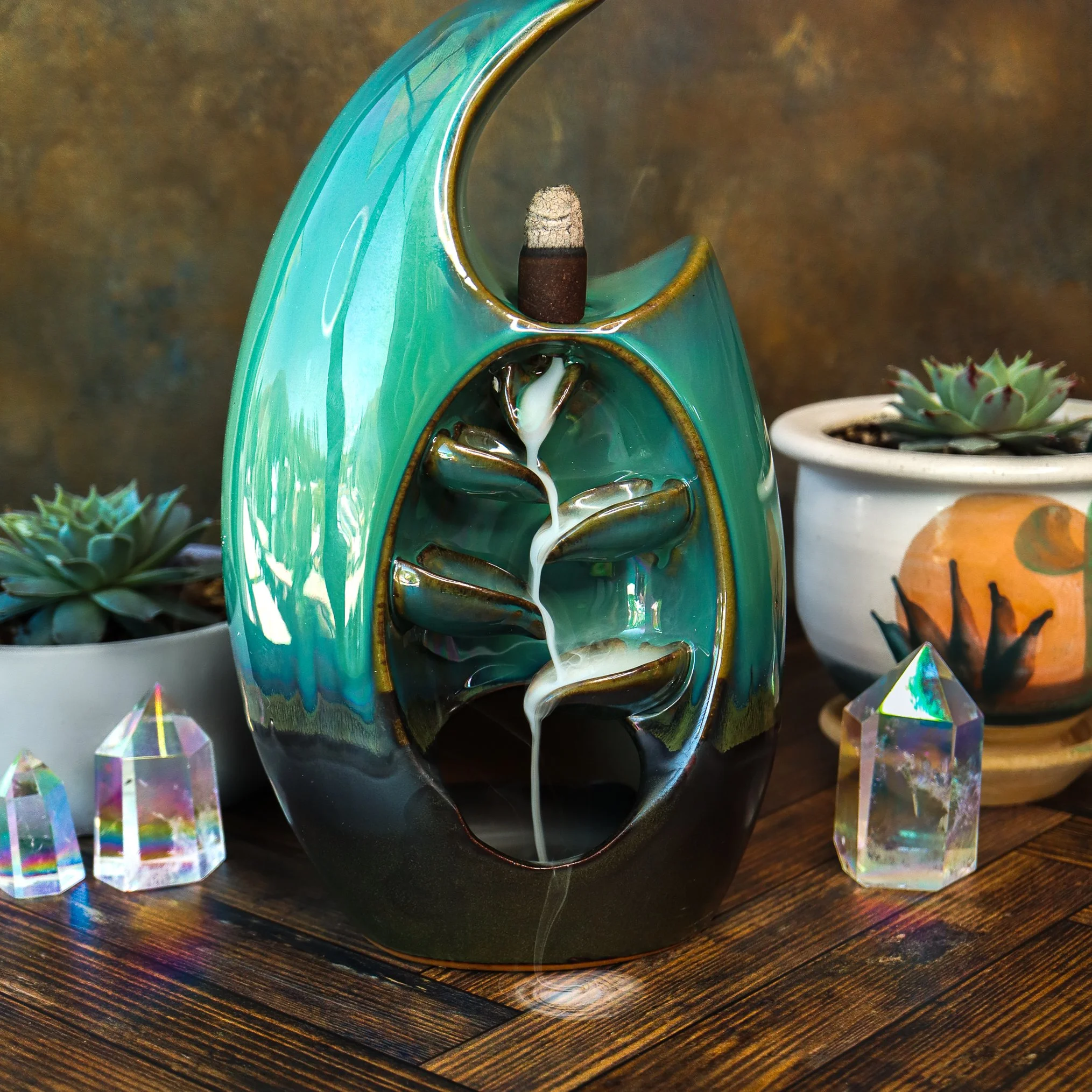A turquoise ceramic incense holder with a spiral design, holding a burnt incense stick. The setup includes a potted succulent plant and crystalline objects on a wooden surface.
