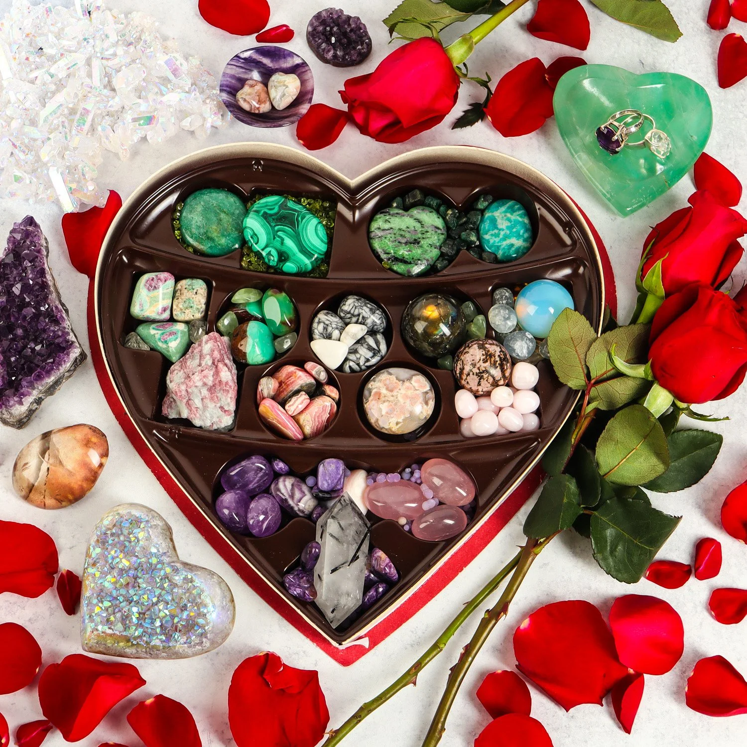 A heart-shaped box filled with various colorful gemstones and crystals, surrounded by red rose petals and roses, with some additional gemstones and crystals scattered around.