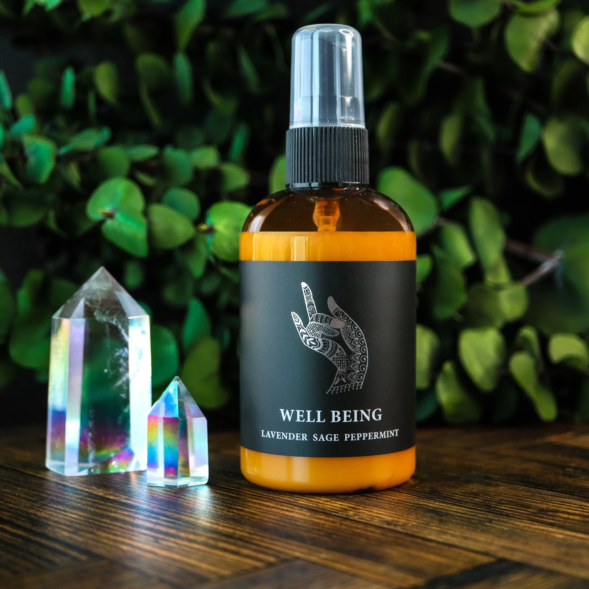 A small amber glass spray bottle with a black label and black spray nozzle, sitting on a wooden surface with a green leafy background.