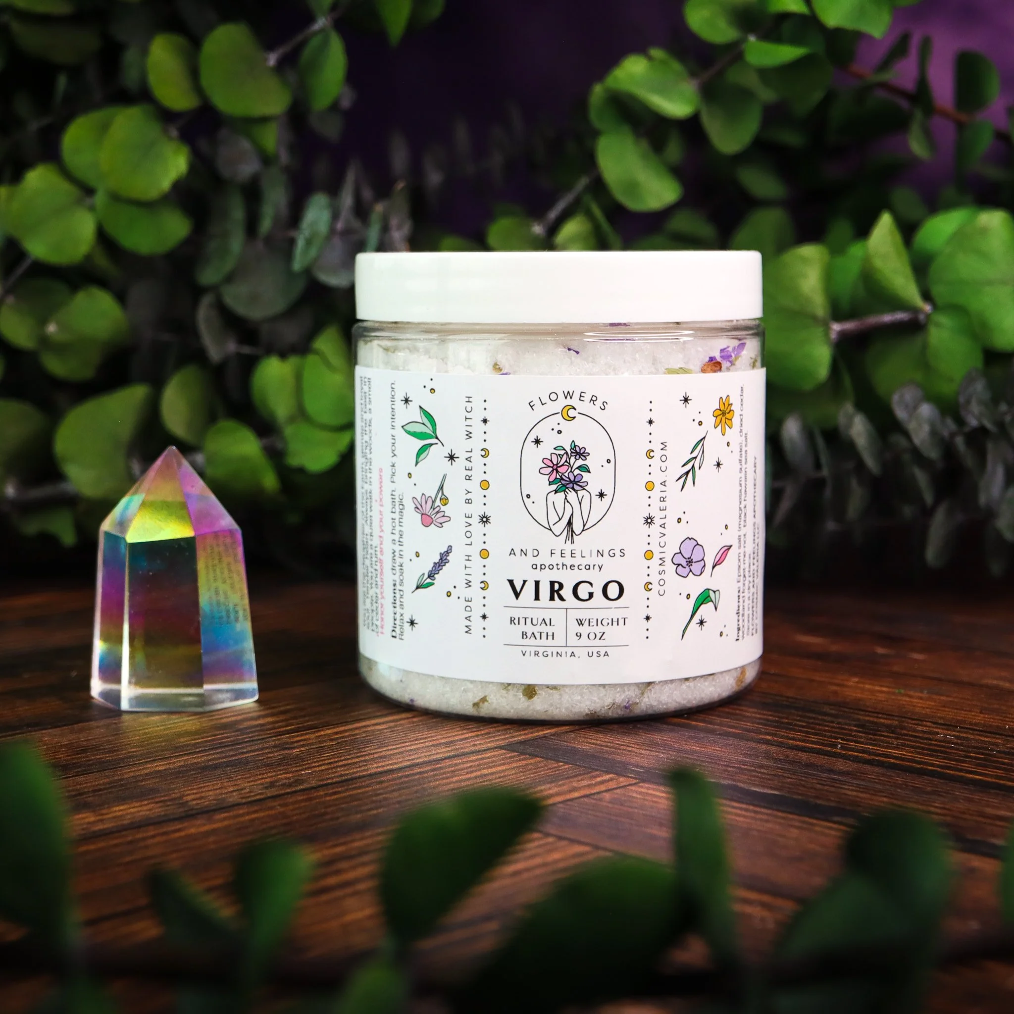 A jar of Virgo-themed bath salts or crystals with a decorative label featuring celestial and floral designs, placed on a wooden surface with greenery in the background.