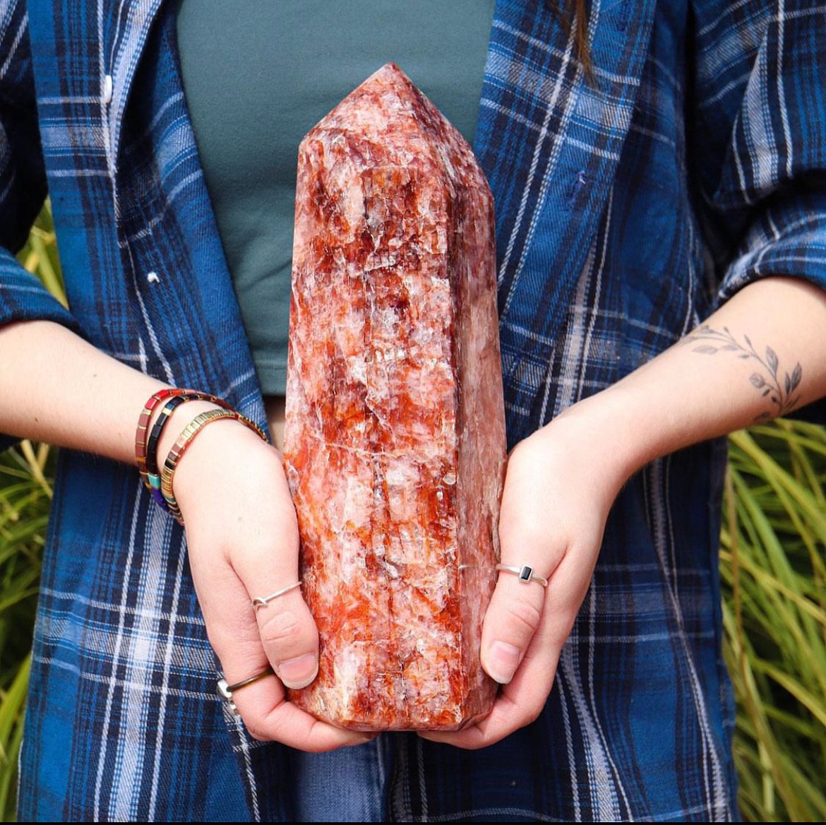 Person holding a large, orange and white salt crystal or mineral specimen. The person is wearing a blue plaid shirt and has tattoos and colorful bracelets on their arms.
