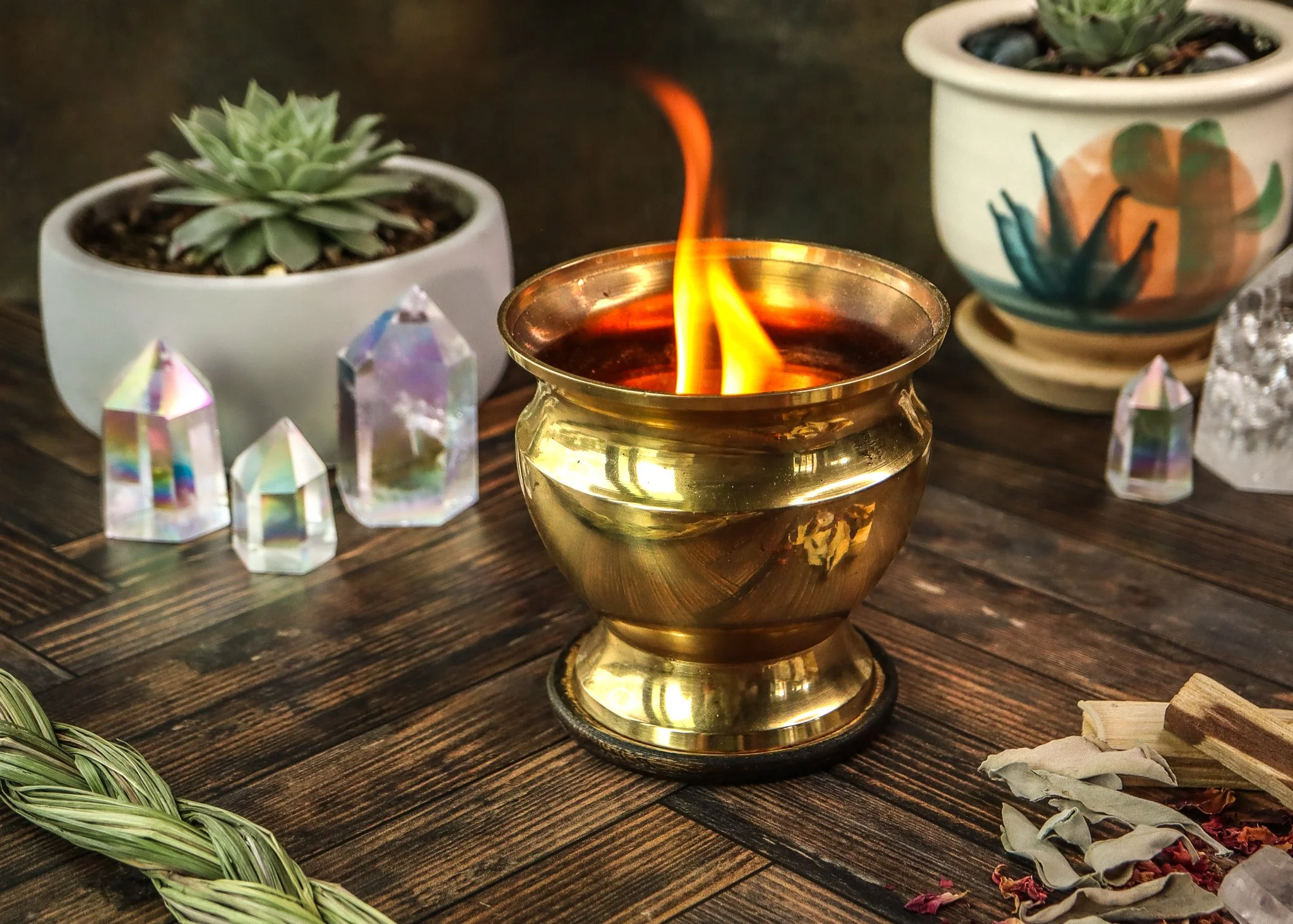 A photograph of a small brass-colored candle holder with a lit flame, placed on a dark wooden surface. Surrounding the candle are two potted succulents, decorative crystals, and dried flower petals or herbs.