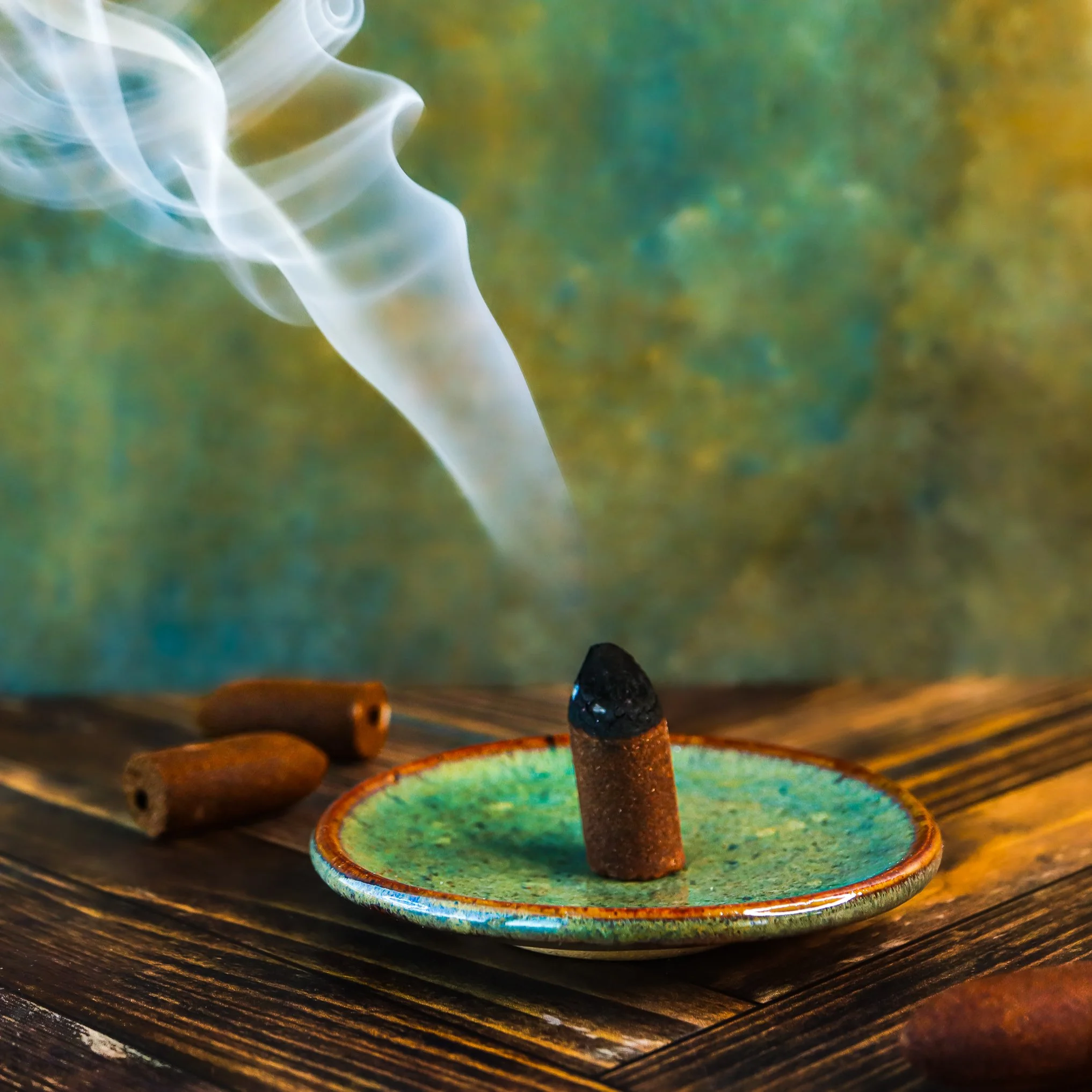 A close-up of small colorful ceramic bowls and candles on a wooden table with smoke rising from a burnt stick, creating a calming and ritualistic atmosphere.