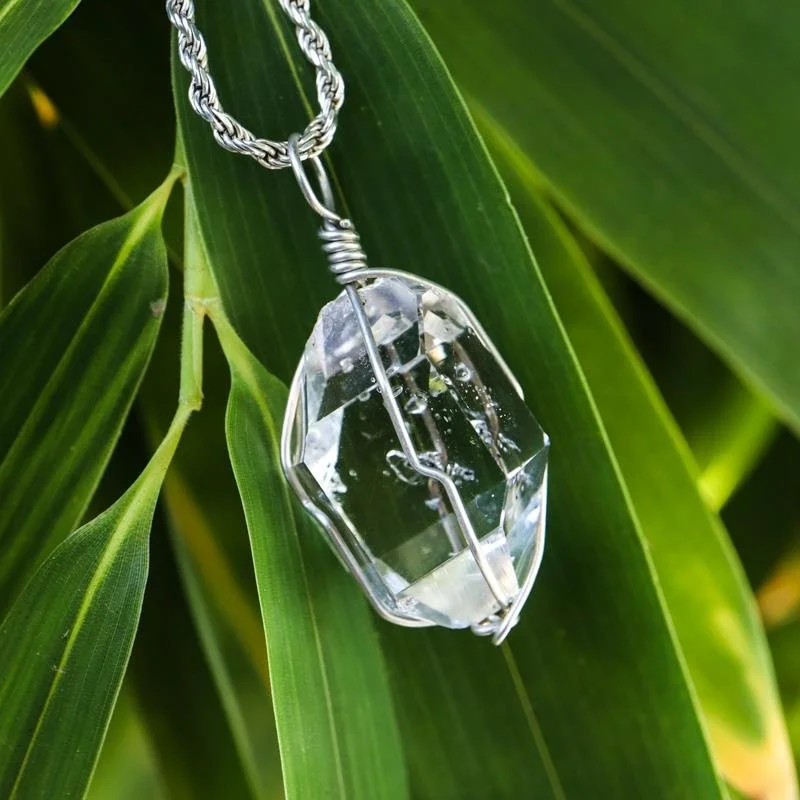 A pendant necklace with a large, clear, faceted crystal hangs from a silver chain, draped over green leaves.