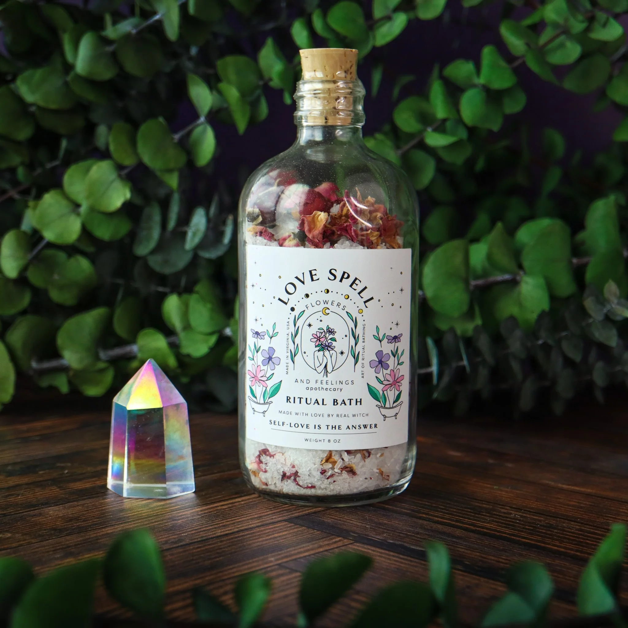 A clear glass bottle filled with dried flower petals and salts, labeled 'Love Spell' Ritual Bath, placed on a wooden surface with green plants in the background and a rainbow prism glass nearby.
