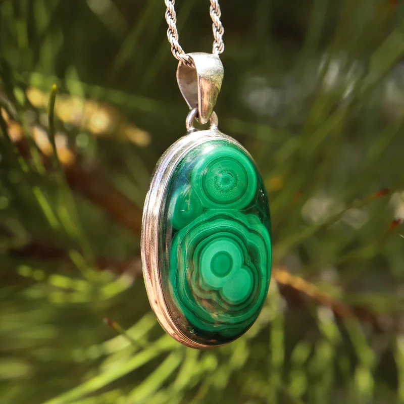 Close-up of a green malachite gemstone pendant on a silver chain, hanging outdoors with blurred green foliage in the background.
