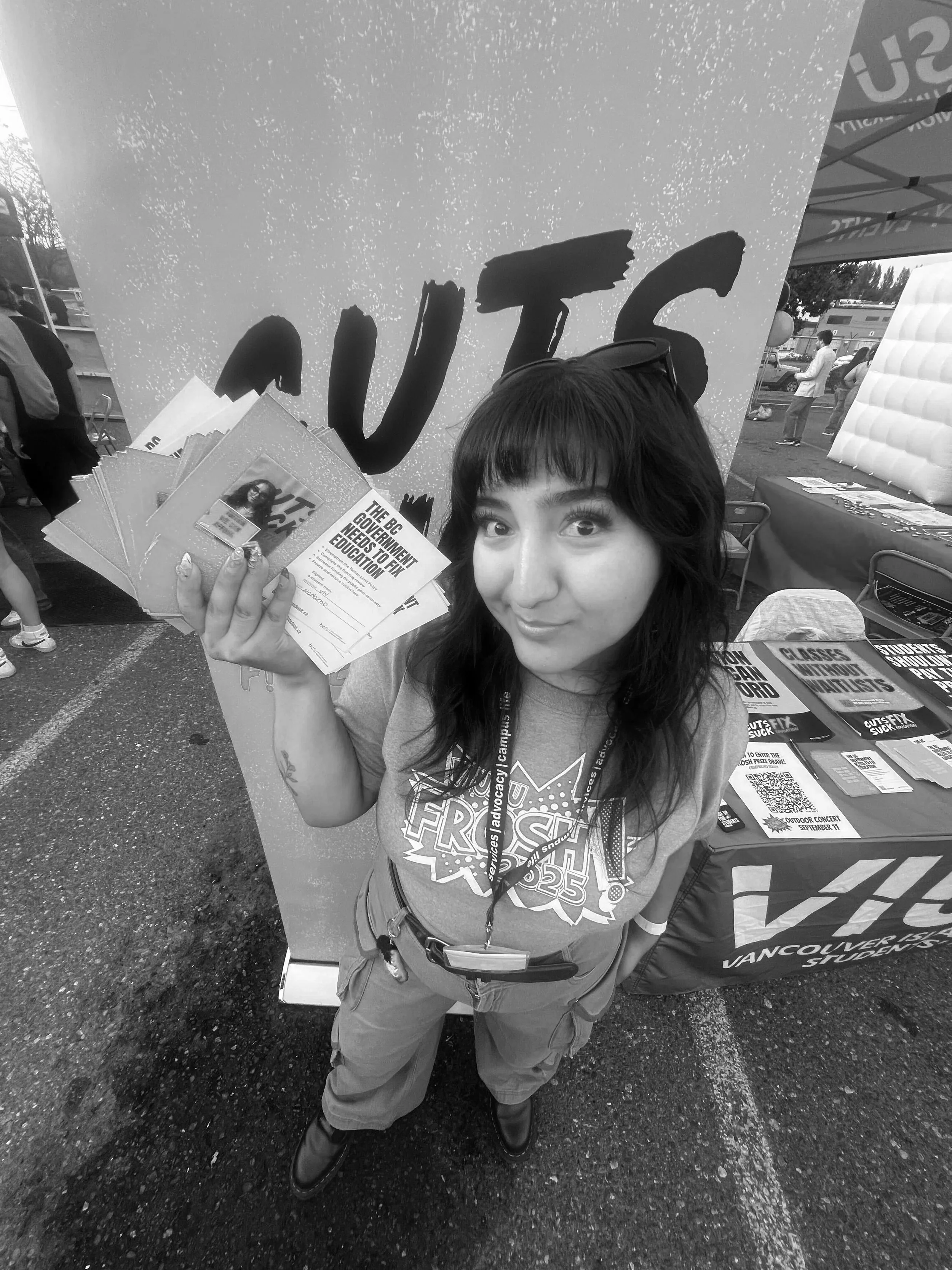 A young woman with dark hair and expressive eyes is smiling and holding a stack of flyers or brochures at an outdoor event. She is wearing a gray T-shirt with graphic text and a lanyard around her neck. Behind her, there is a booth with a large sign and a table displaying more flyers and materials. Other people are visible in the background at the event.